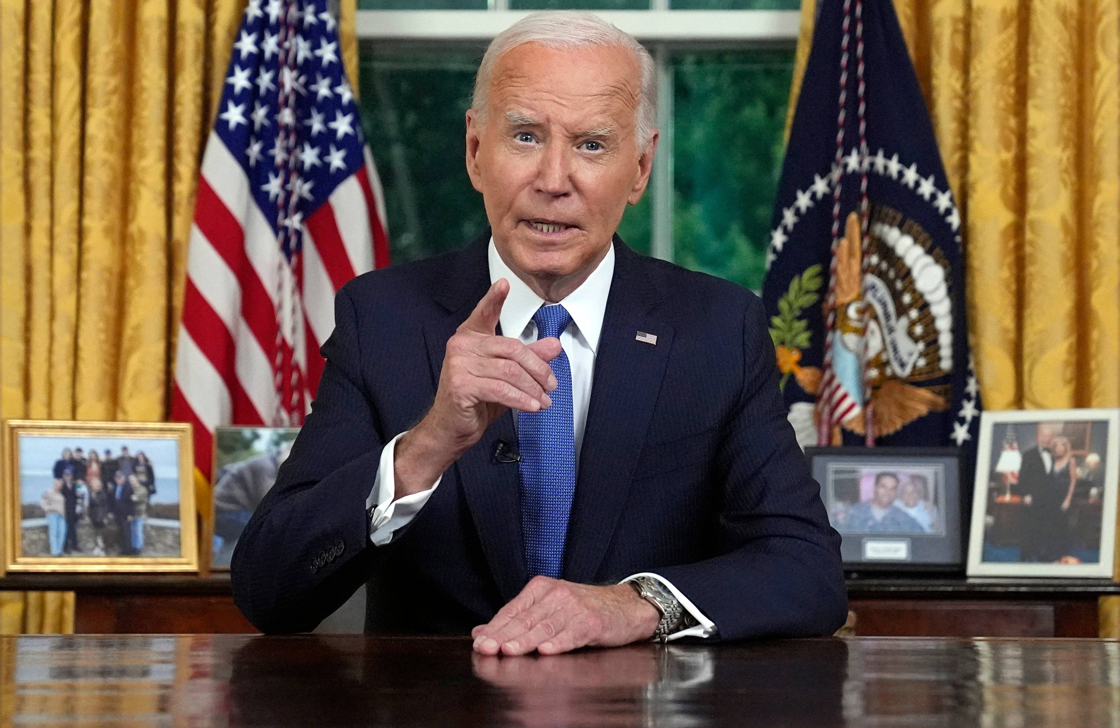 Prezydent Joe Biden ubrany w ciemny garnitur i błękitny krawat przemawia siedząc przy biurku w gabinecie owalnym w Białym Domu. Za jego plecami wisi amerykańska flaga oraz sztandar prezydenta USA.