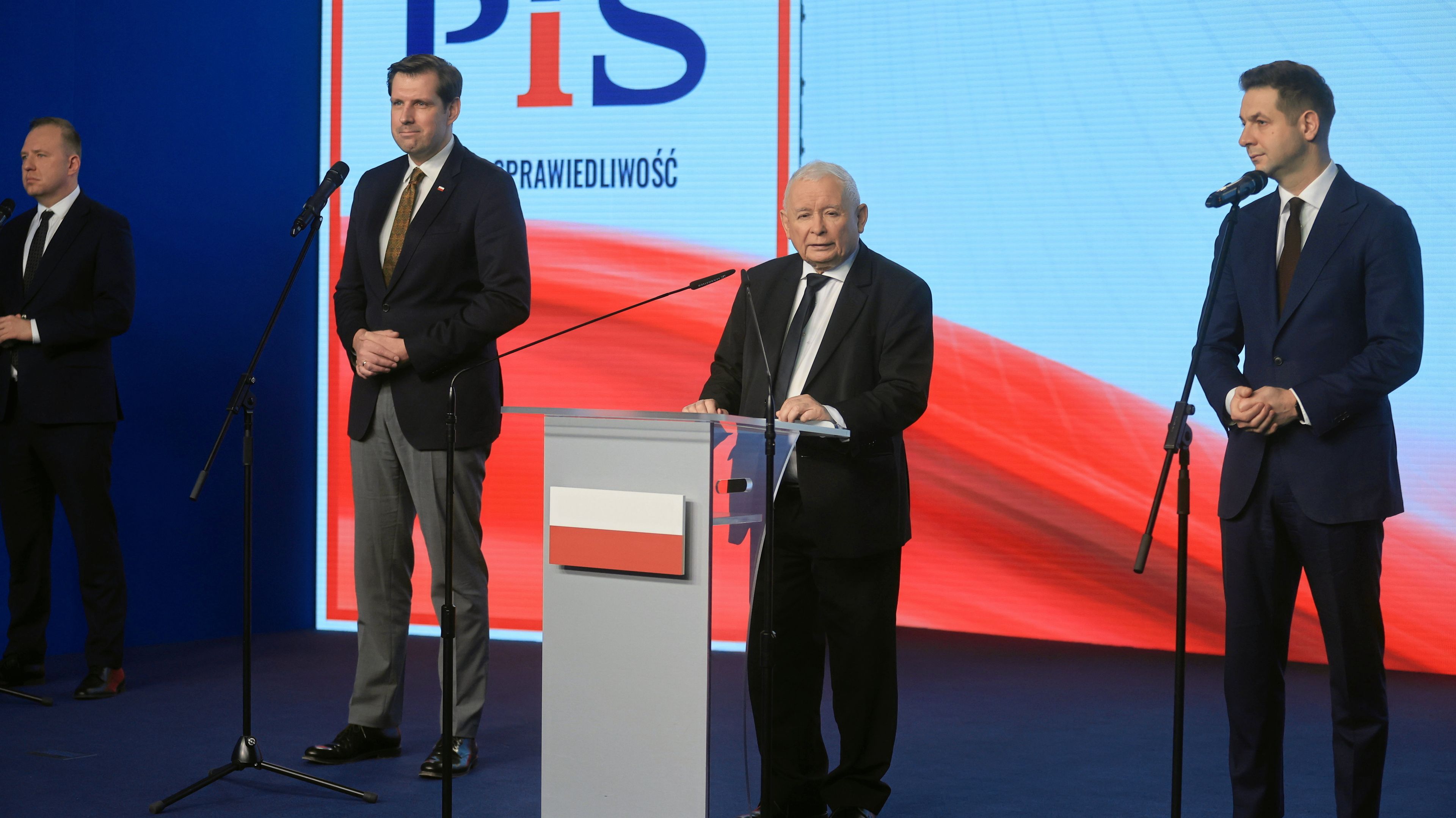Od lewej: Tobiasz Bocheński, Jarosław Kaczyński, Patryk Jaki, w tle logo PiS, z przodu mównica z flagą Polski