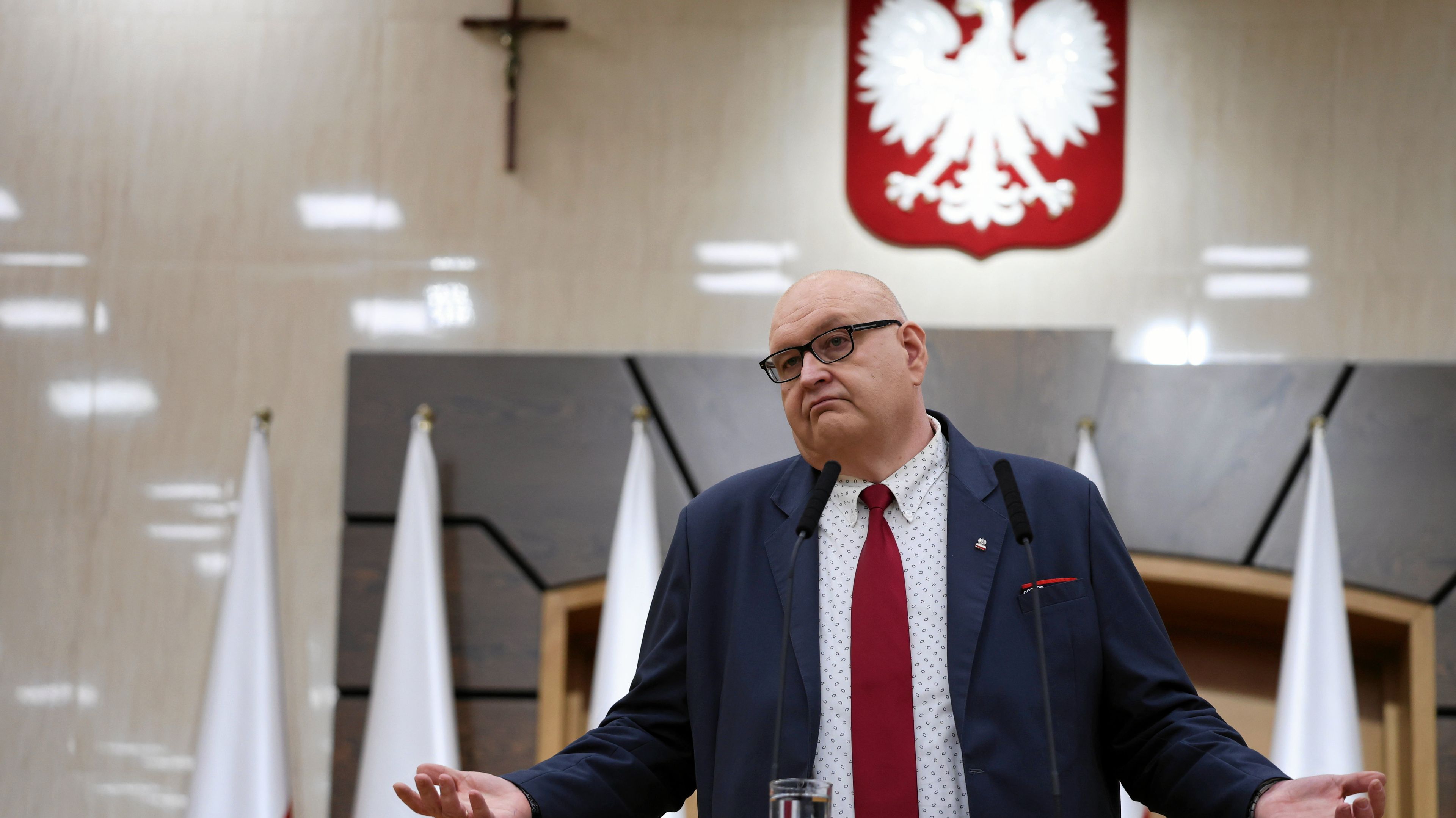 Otyłu mężczyzna w okulrach, łysawy, stoi i przemawia rozkładając ręce