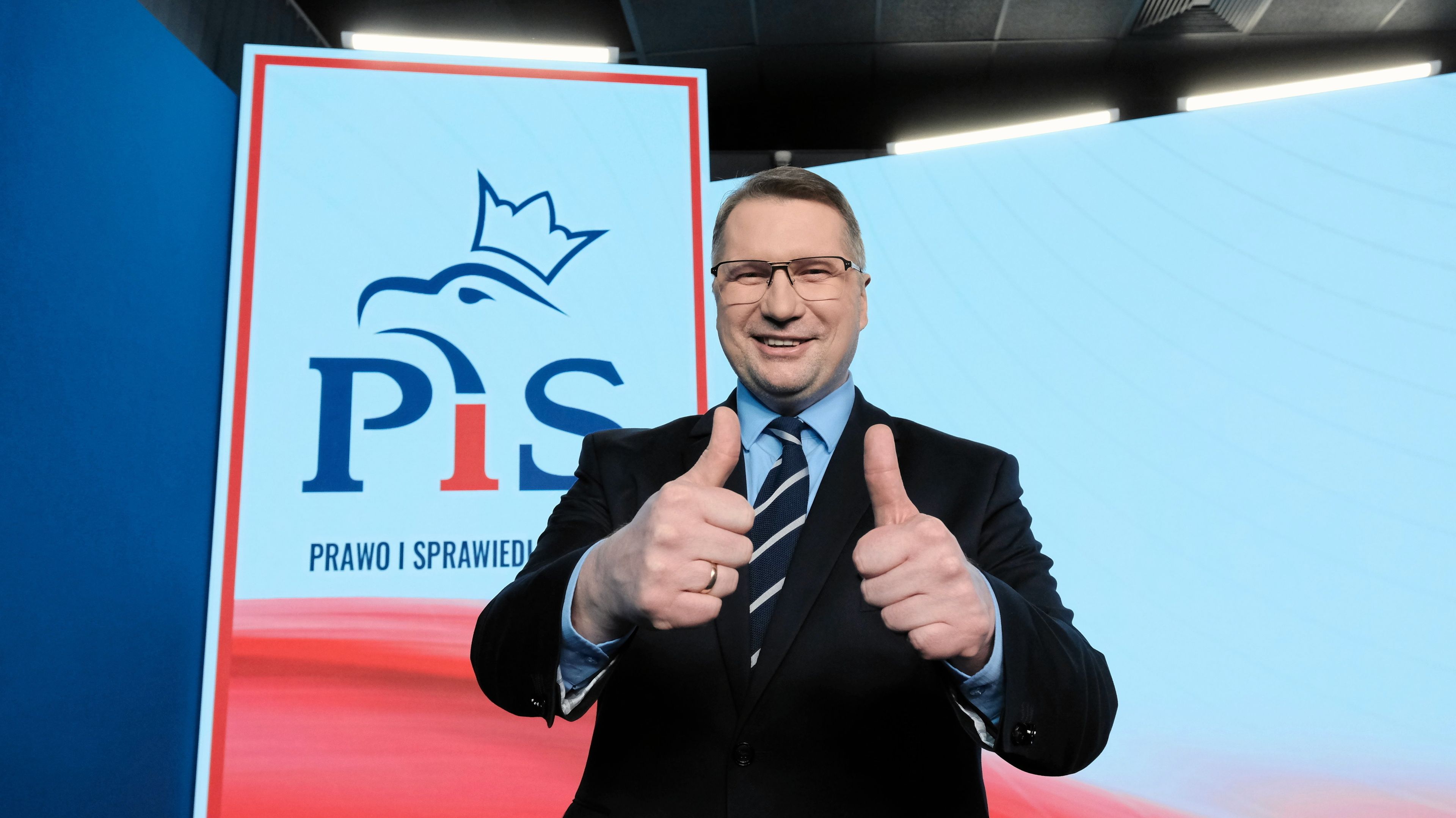 Przemysław Czarnek pokazuje gest dwóch kciuków w górę na tle logo PiS