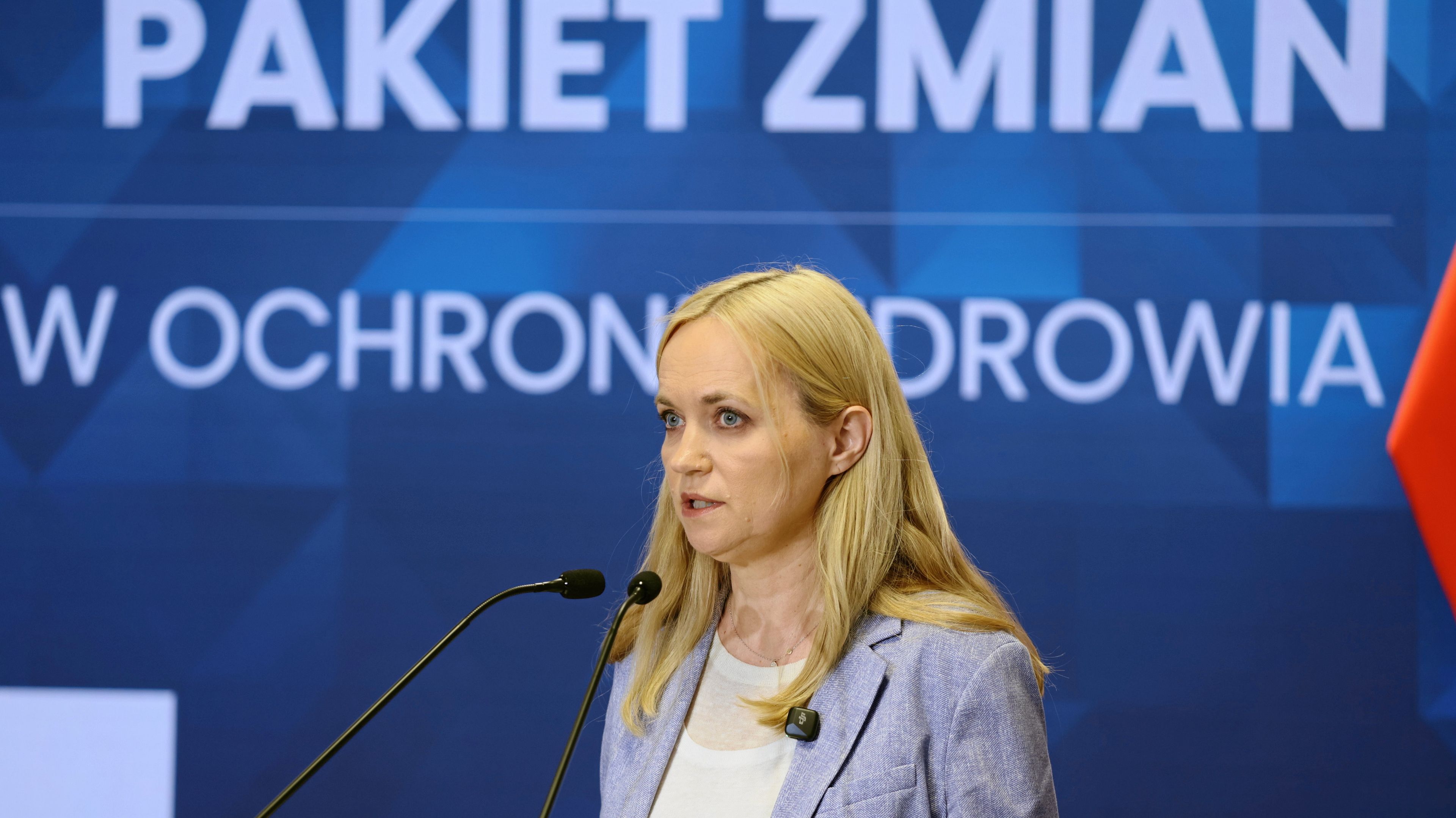 Młoda kobieta z długimi blond głosami przemawia na tle wielkiego napisu Pakiet zmian w ochronie zdrowia