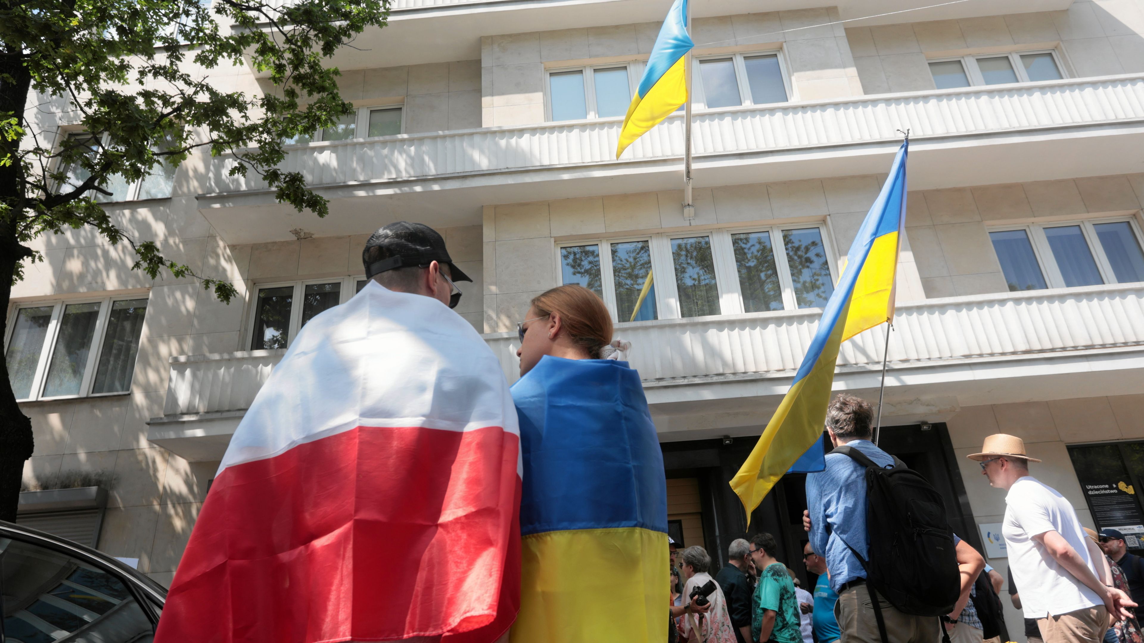 dwie osoby stoją obok siebie. Jedna jest okryta biało-czerwoną flagą, druga - flagą Ukrainy