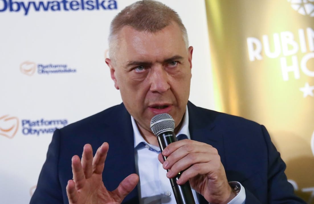 Roman Giertych na konferencji prasowej. Przemawia do mikrofonu, który trzyma w ręce. Ubrany w ciemną marynarkę i rozpiętą pod szyją jasną koszulę.