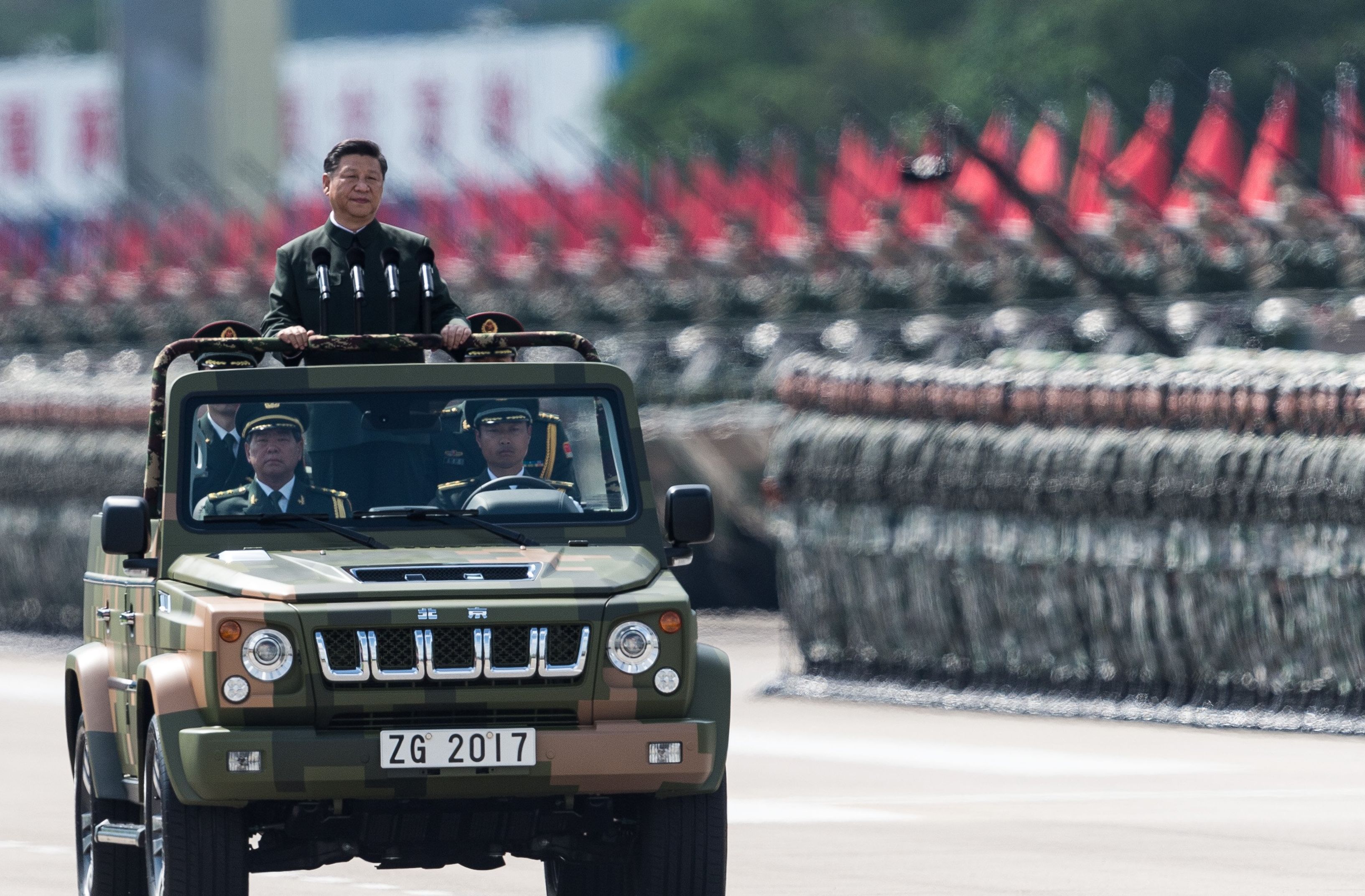 Przewodniczący Chińskiej Republiki Ludowej Xi Jinping podczas defilady wojskowej. Chiński przywódca jest wieziony wojskowym samochodem tak, że wystaje ponad jego dach i może pozdrawiać żołnierzy. W tle równe zastępy wojska i uzbrojenia.