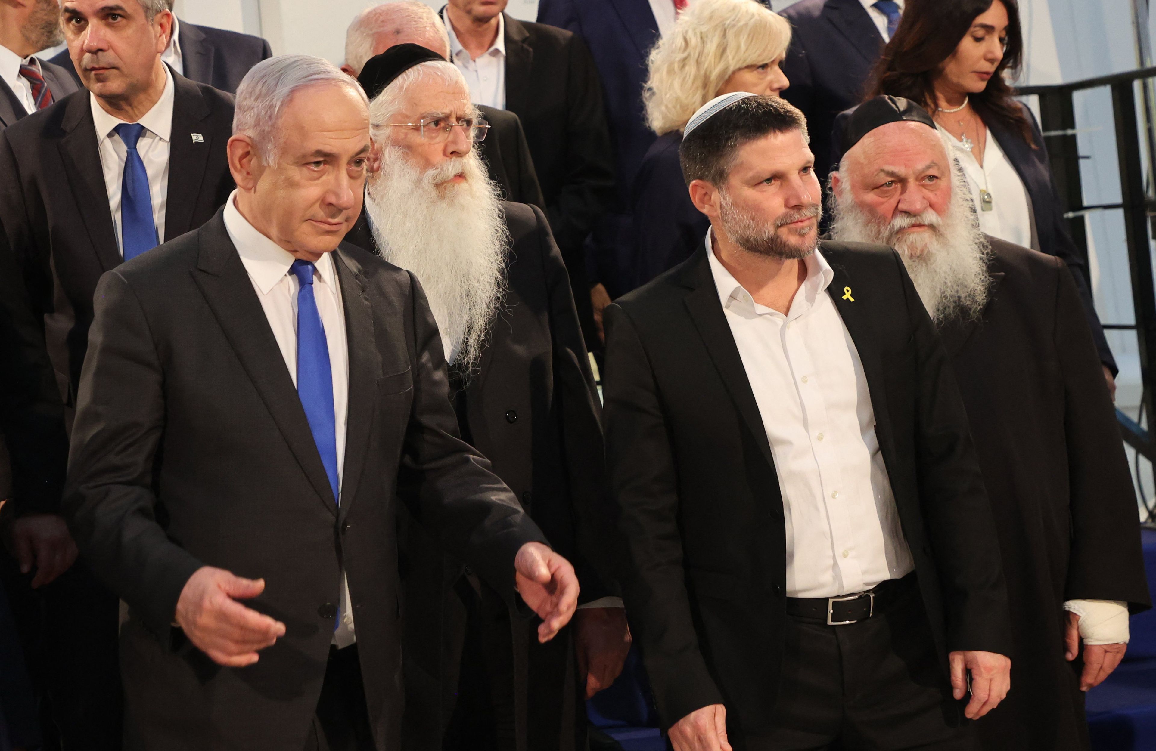 Grupa starszych mężczyzn w czarnych garniturach, w widocznych na głowach jarmułkach idzie korytarzem na spotkanie.