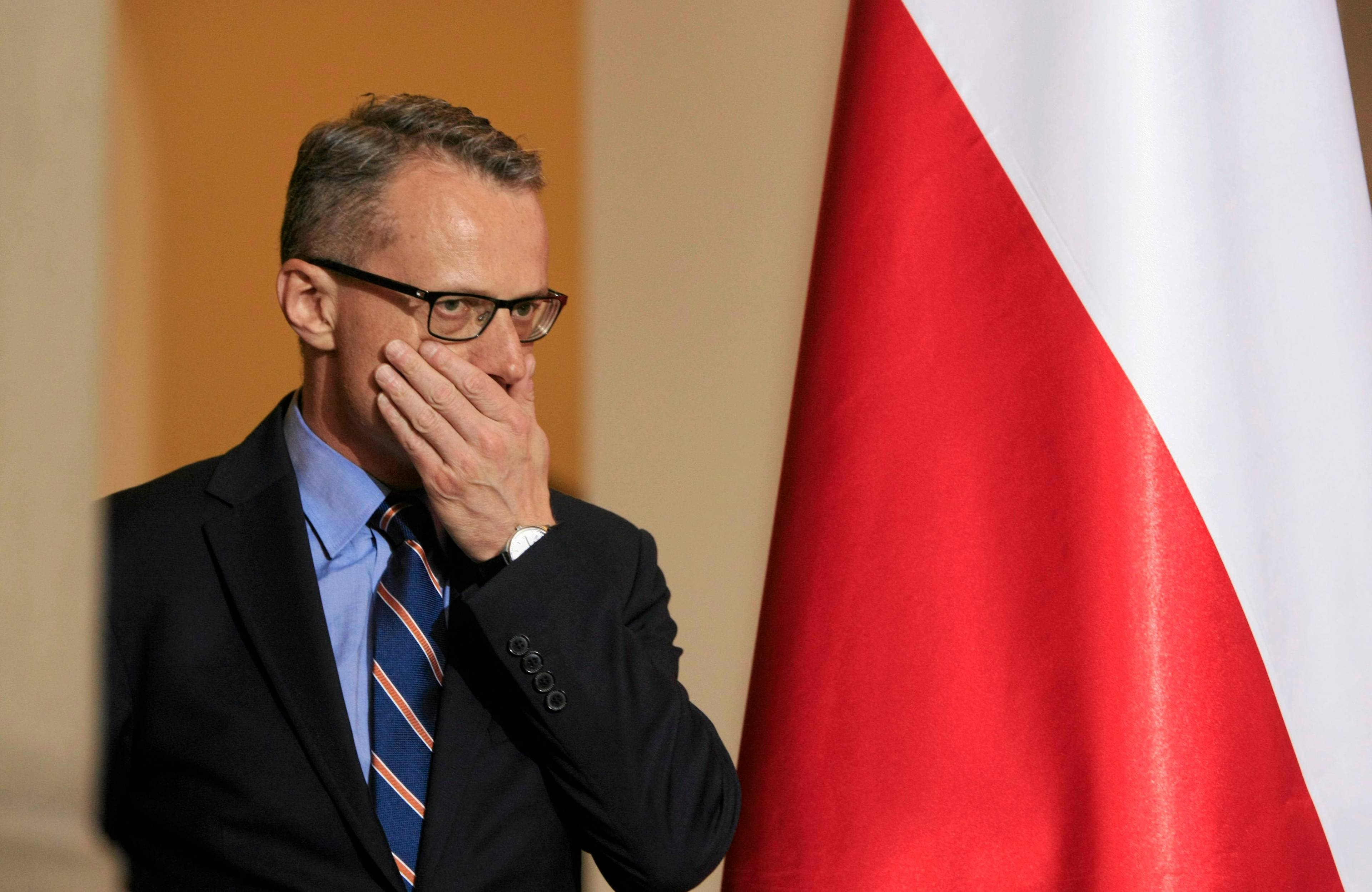 40-letni mężczyzna w okularach i garniturze zasłania twarz ręką, w tle polska flaga