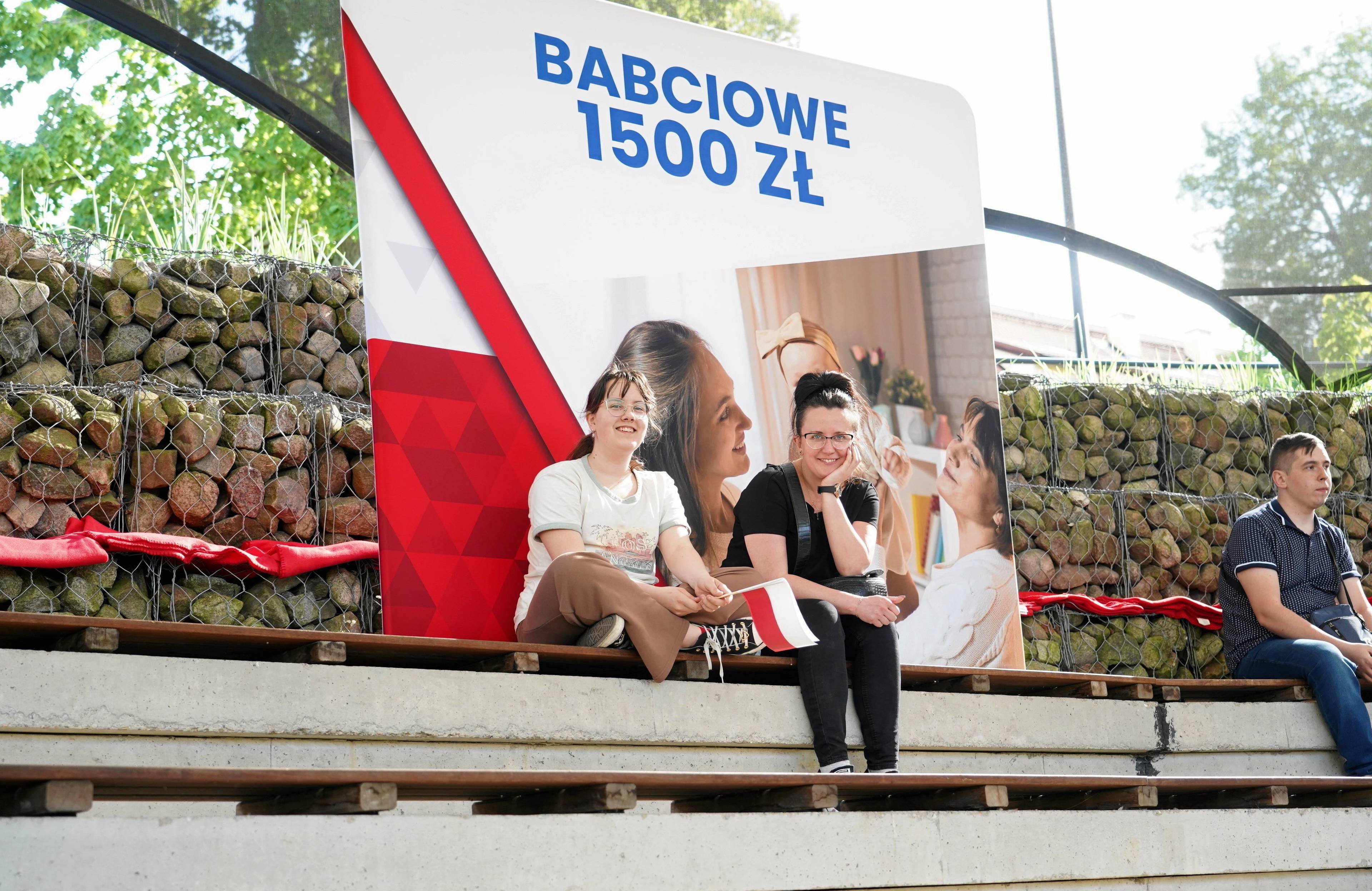 Dwie dziewczyny siedzą na tle plakatu z transparentem "Babciowe 1500 zł"