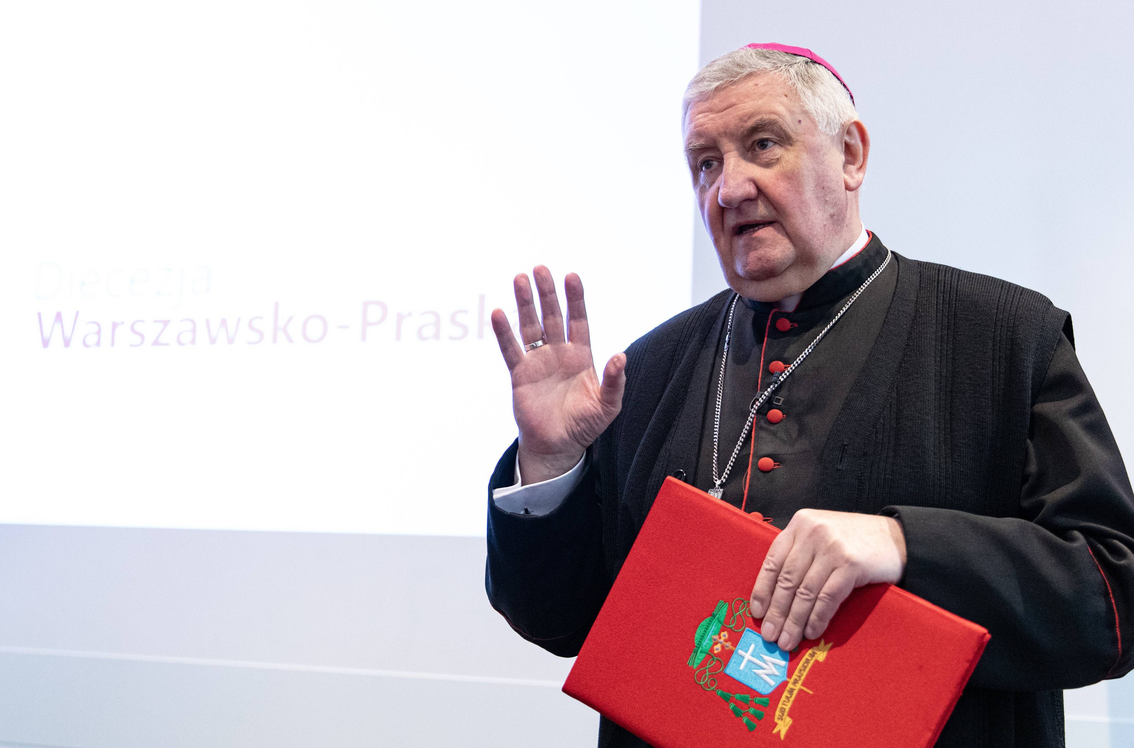 Biskup Romuald Kamiński stoi z czerwoną teczką w ręku. Drugą rękę trzyma w górze