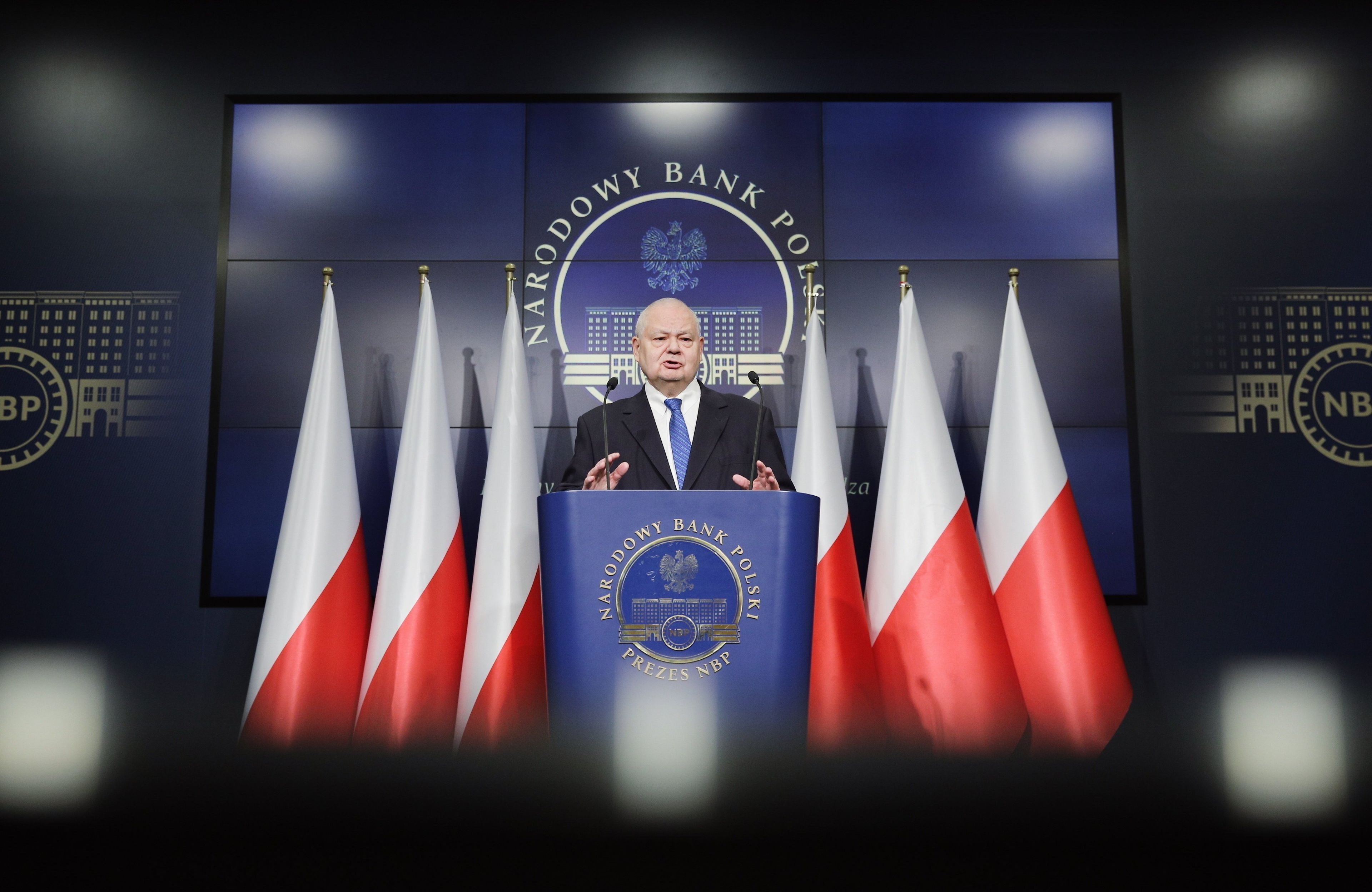 prezes NBP Adam Glapiński na tle flag polski przemawia do mikrofonów
