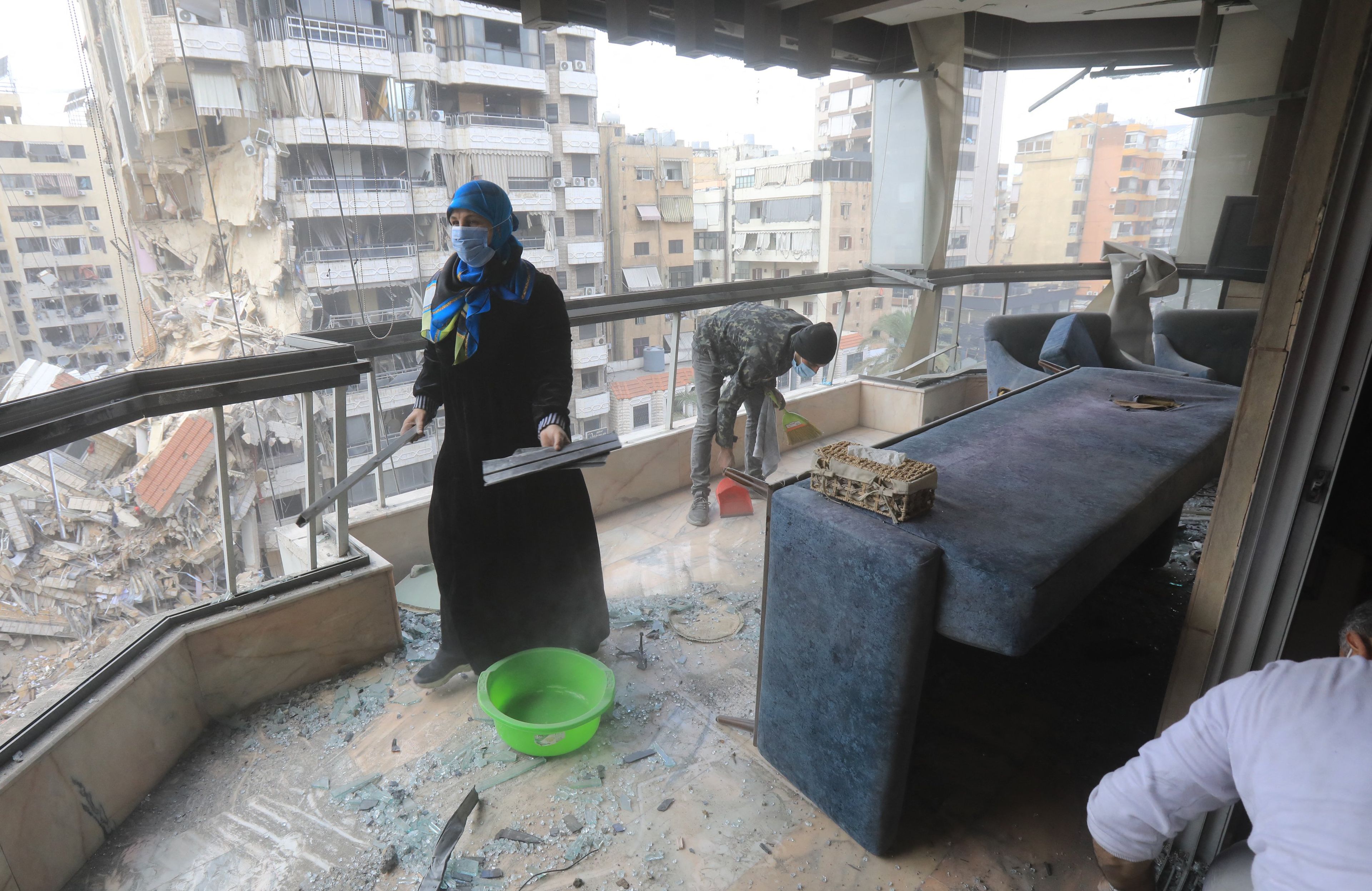 27.11.2024, Bejrut, Liban. Cywile wracają do swoich domów po ogłoszeniu zawieszenia broni między Izraelem a Hezbollahem. Na zdjęciu kobieta w czarnej sukni sprząta kawałki rozbitego szkła z podłogi swojego mieszkania. Za oknami rozpościera się widok na zniszczone bloki mieszkalne.