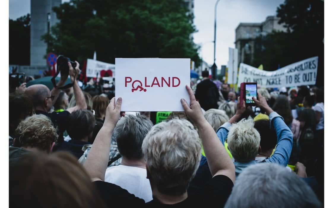 Zgromadzenie. Człowiek trzyma plakat z napisem "Poland", w którym "O" to wywrocony wózek inwalidzki