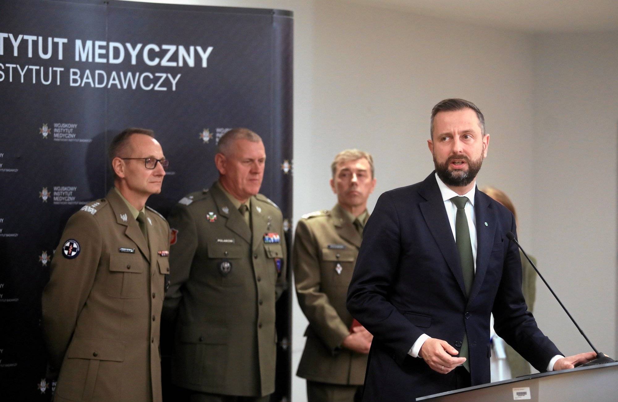 Kosiniak-Kamysz podczas konferencji w Wojskowym Instytucie Medycznym. Przemawia zza podium, z tyłu za nim troje wojskowych w mundurach.