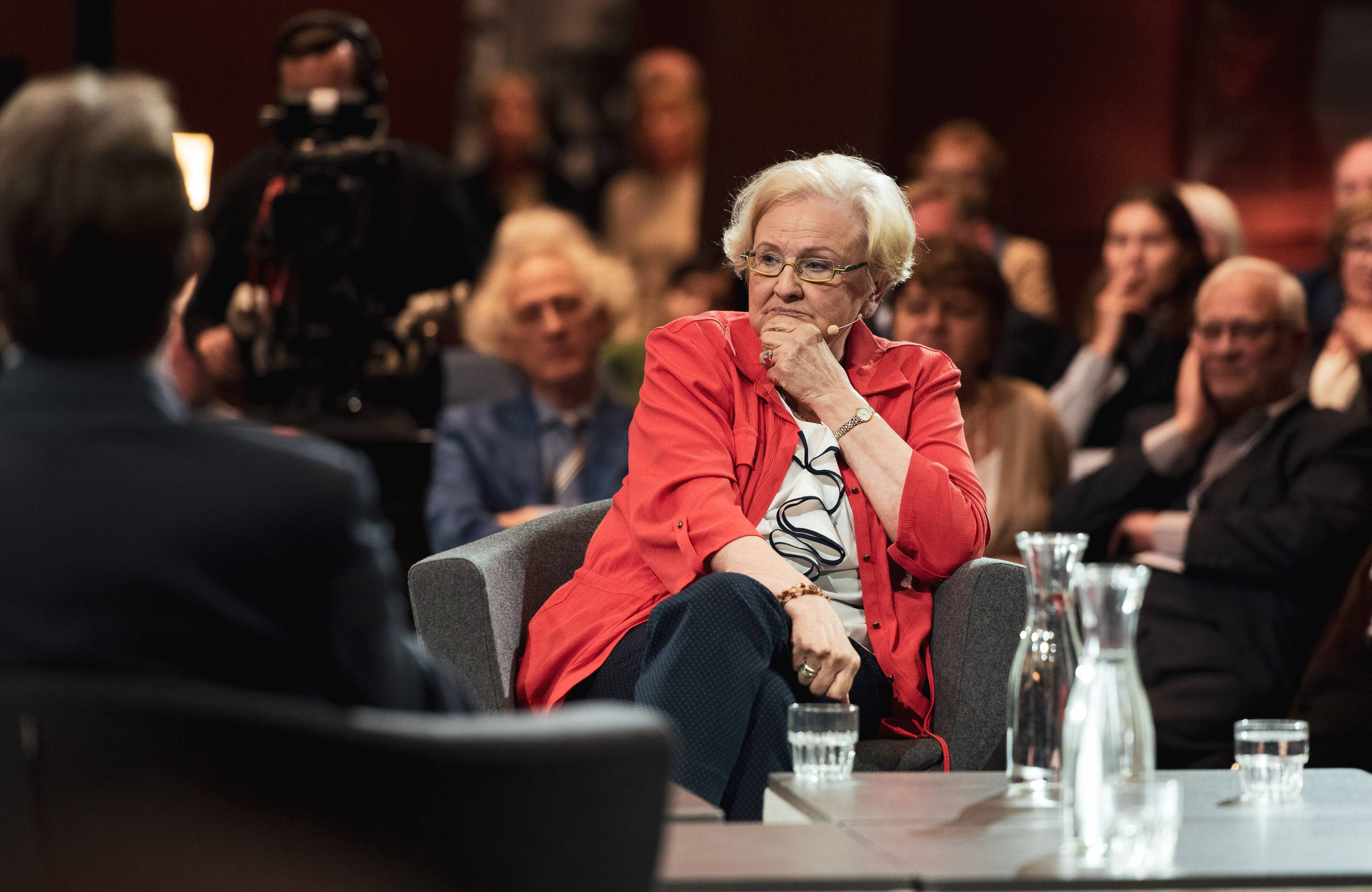 Kobieta w jasno-czerwonym żakiecie siedzi w fotelu, z nią publiczność