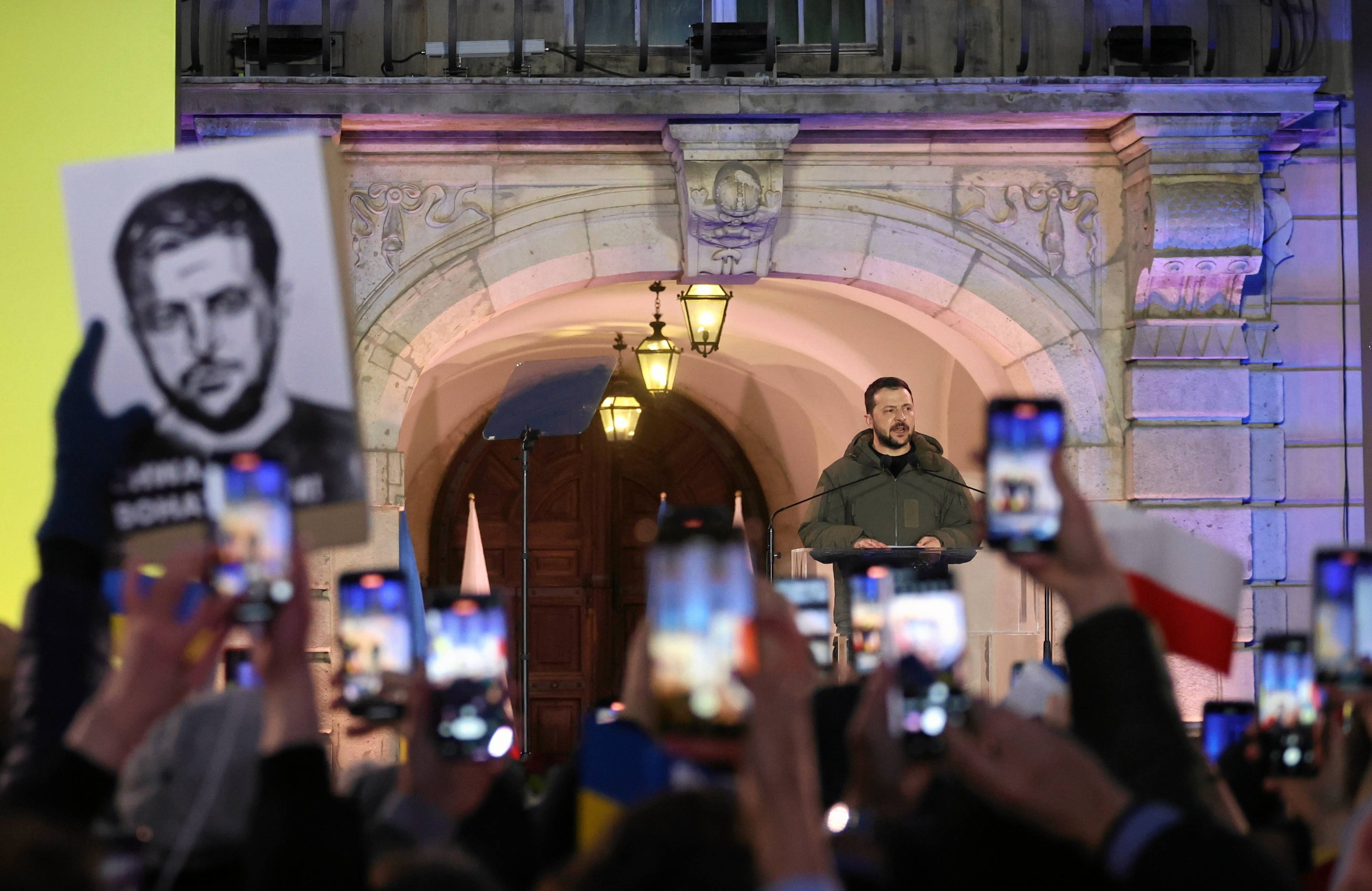 Dziedziniec Zamku Królewskiego, wieczór, oświetlenie, z podwyższenia przemawia mężczyzna, to prezydent Ukrainy Wołodymyr Zełenski