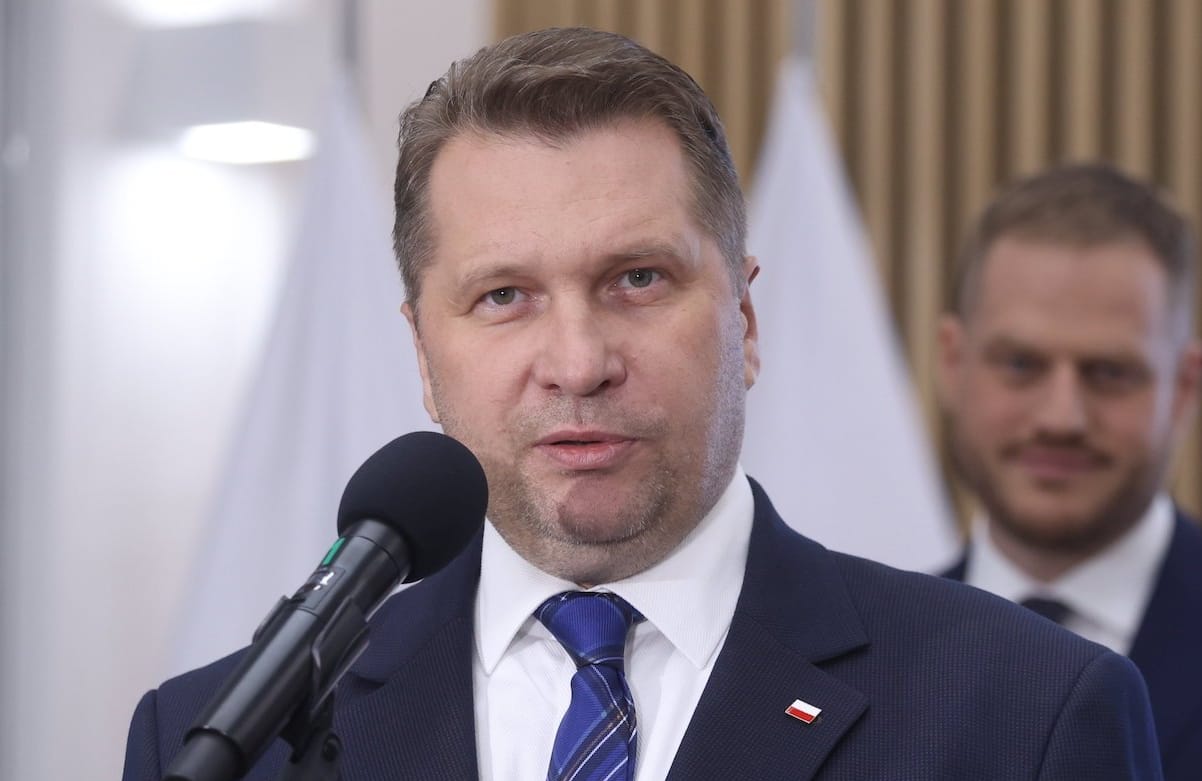 Minister Przemysław Czarnek na konferencji prasowej przemawia do mikrofonu, ubrany w granatowy garnitur, białą koszulę i niebieski krawat. W klapie marynarki przypinka z flagą Polski. W tle stoi minister cyfryzacji Janusz Cieszyński, ale jest nieostry.