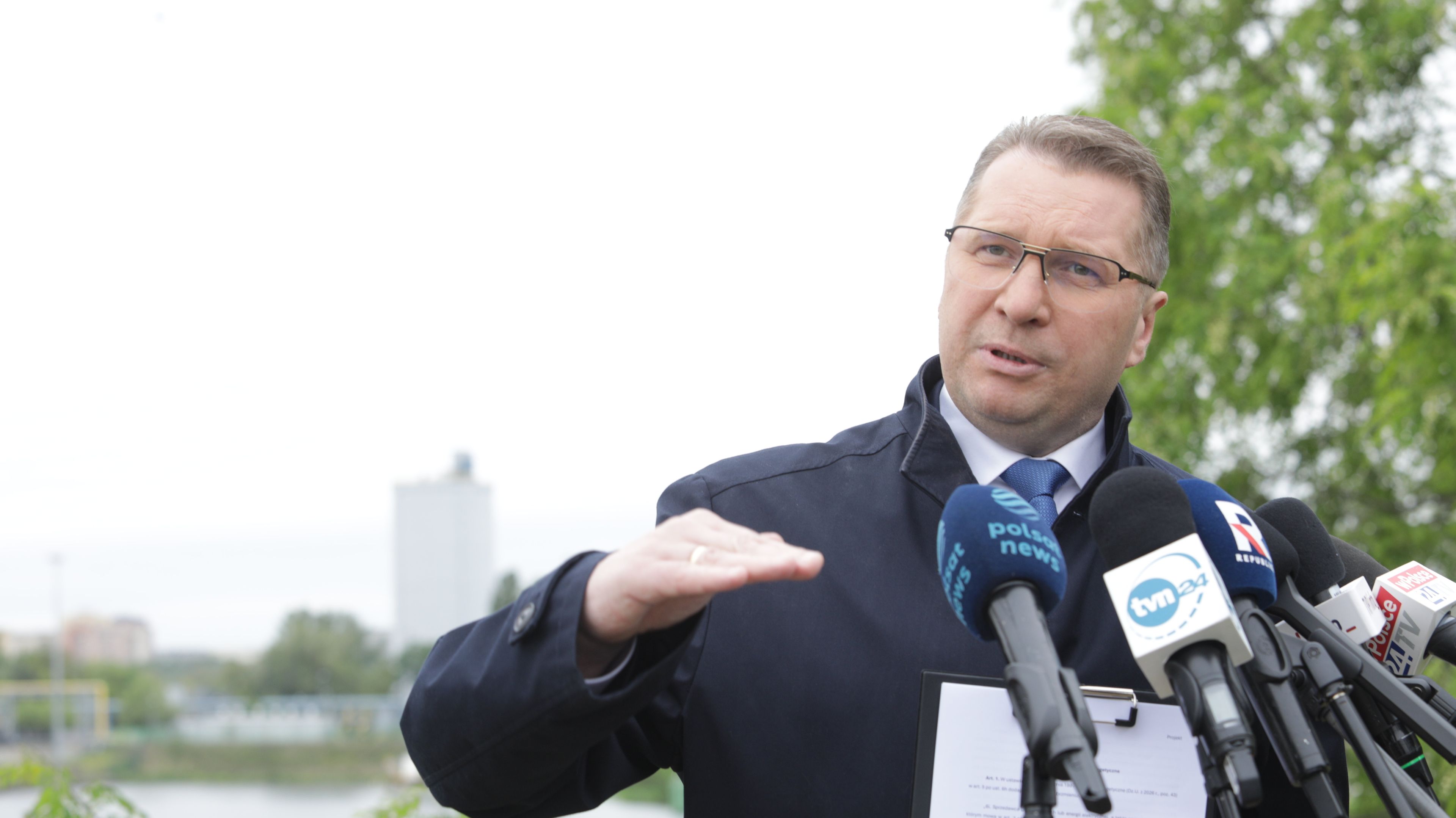 Mężczyzna w okularach tłumaczy coś dziennikarzom pokazując jakiś poziom dłonią