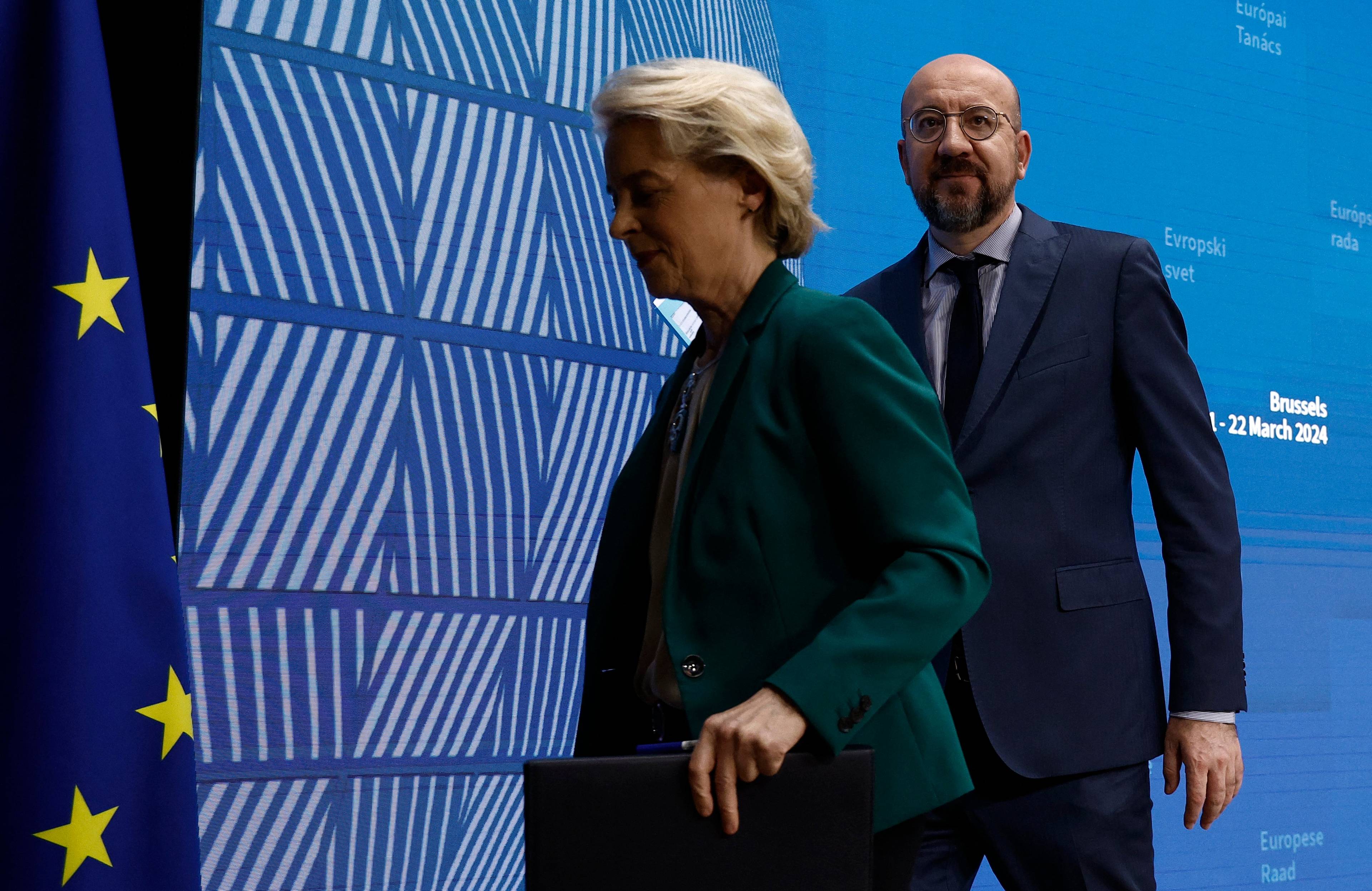 Kobieta w zielonej garsonce i mężczyzna w garniturze przechodzą obok niebieskiej ścianki i flagi Unii Europejskiej