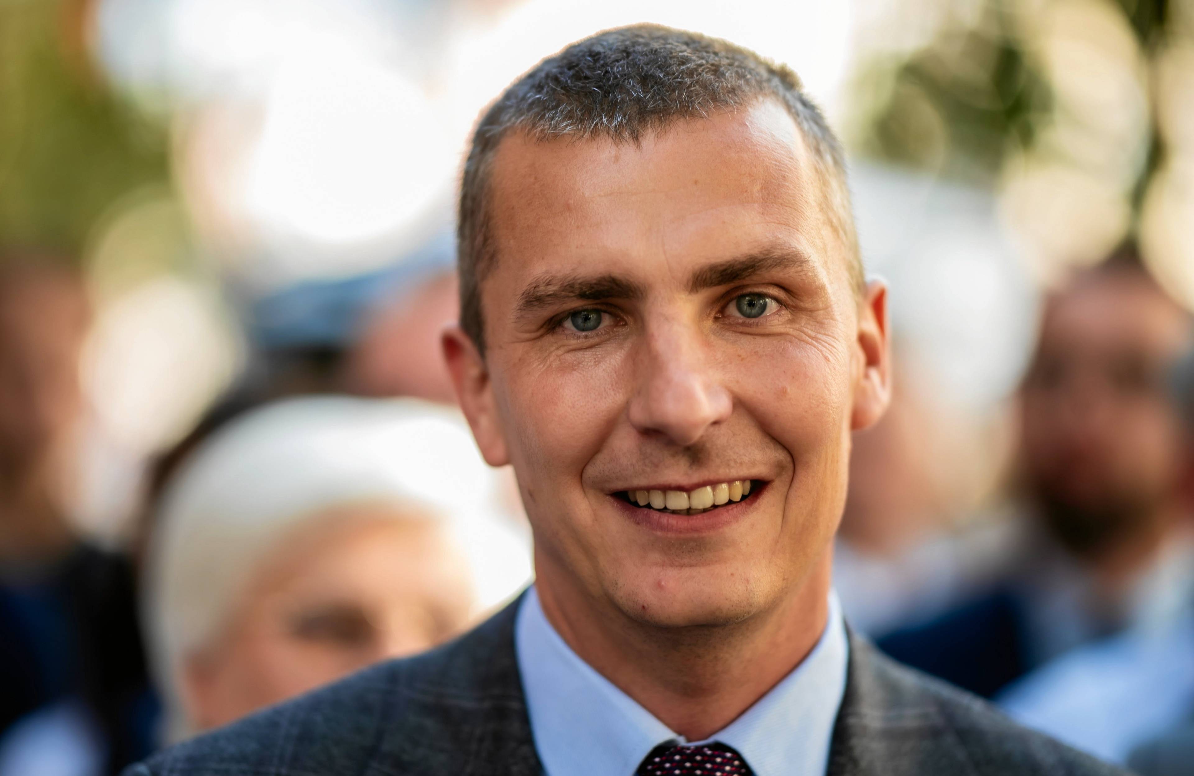 uśmiechnięty 30-latek o szczupłej twarzy, w garniturze i krawacie