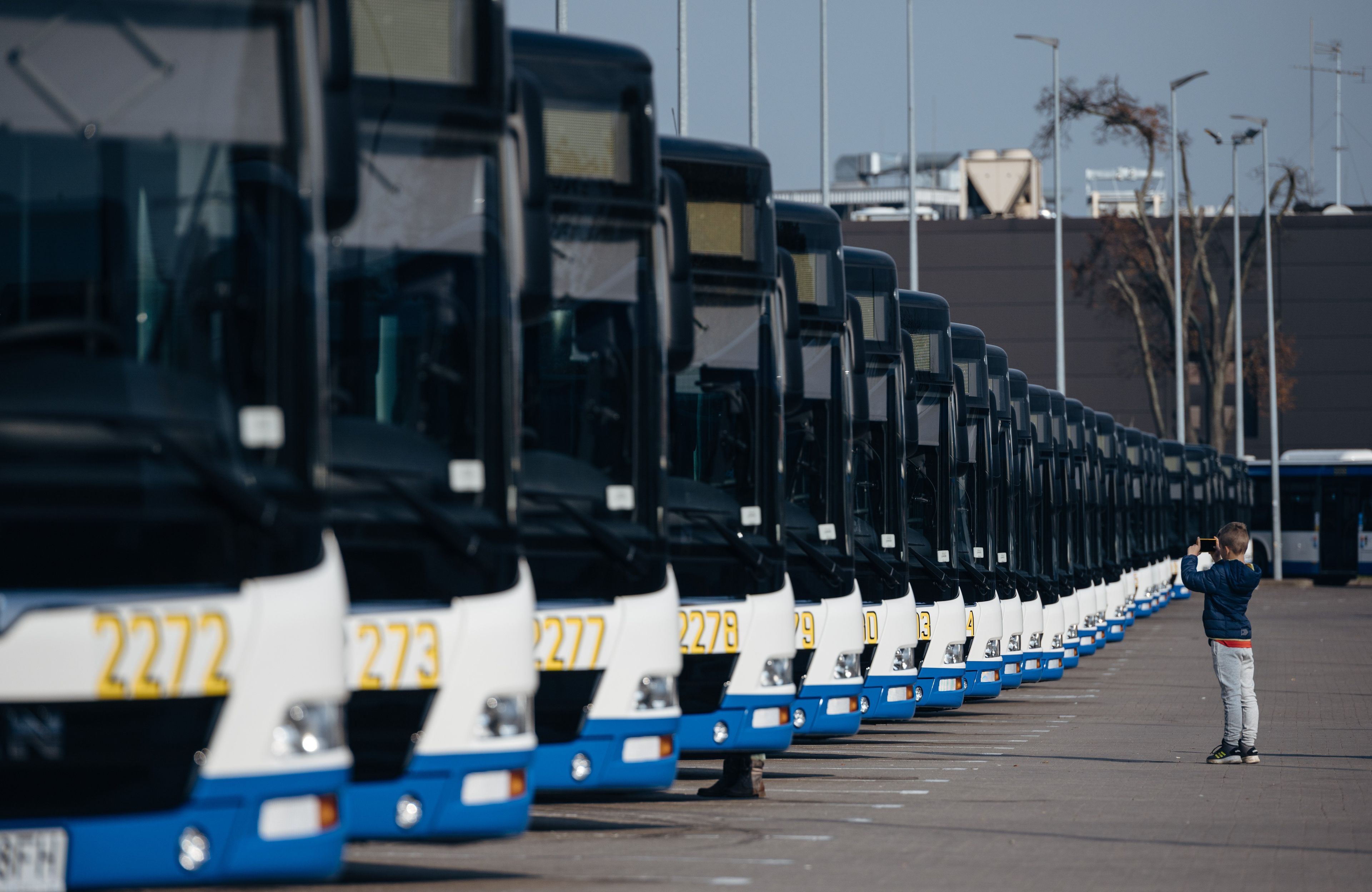 Autobusy miejskie stoją na parkingu. Przed nimi stoi mały chłopiec