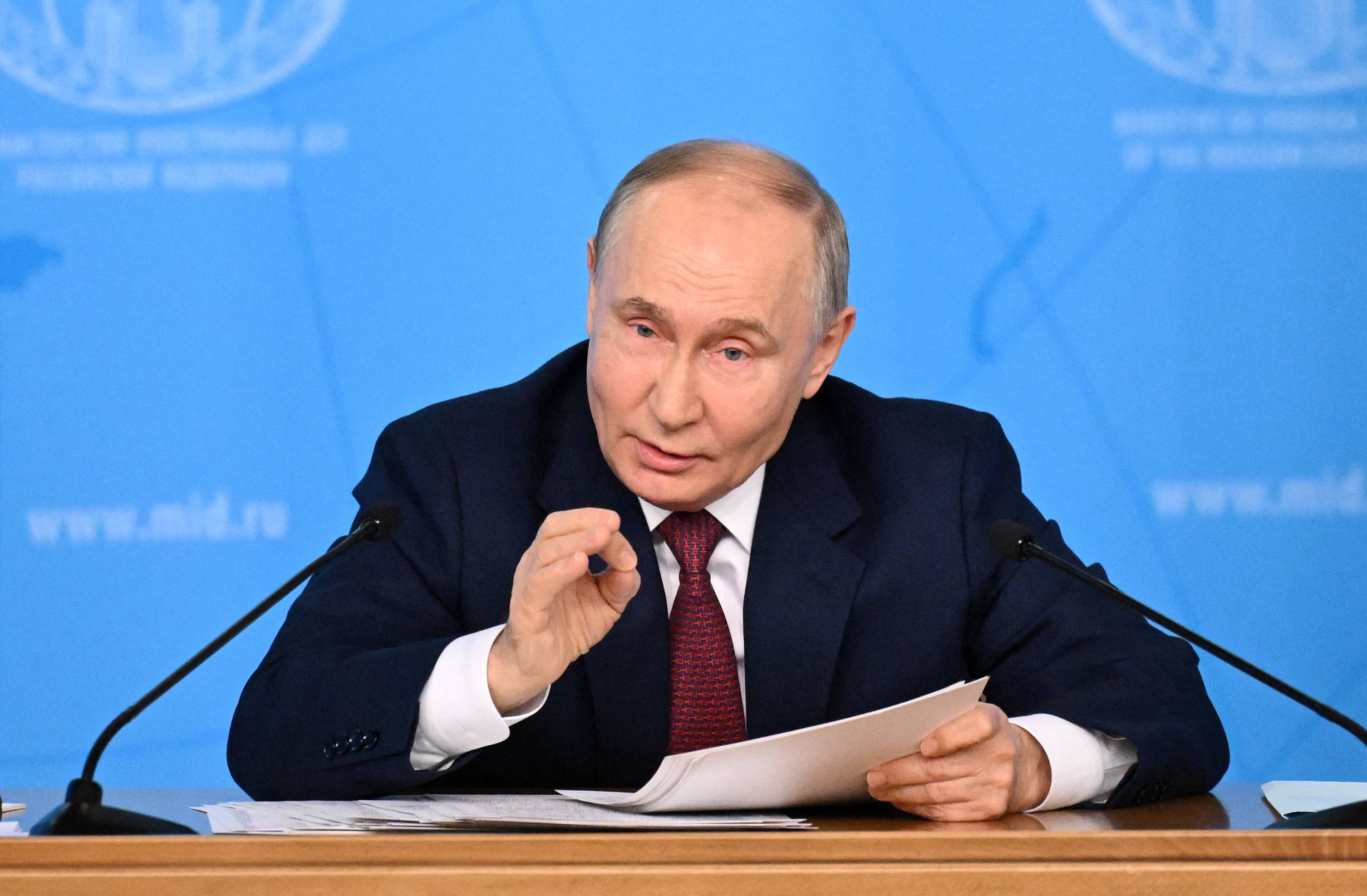 Władimir Putin. Łysiejący, krępy mężczyzna w garniturze i białej koszuli, siedzi przy stole, gestykuluje jedną dłonią.