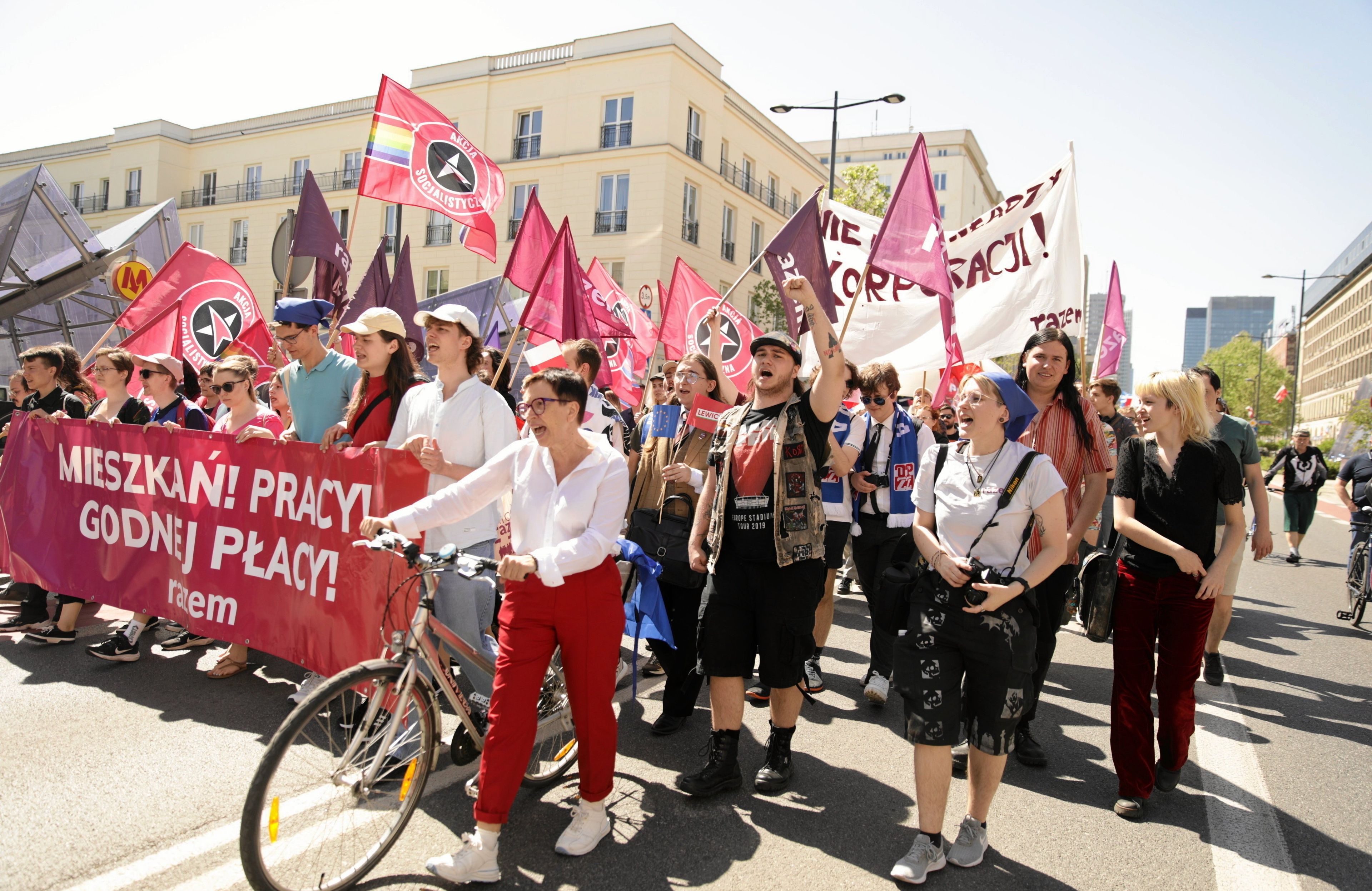 Grupa ludzi z flagami idzie środkiem ulicy w pochodzie pierwszomajowym. Niosą flagi i transparenty, transparent na pierwszym planie głosi "Mieszkań, pracy, godnej płacy". To transparent partii Razem, bo widnieje na nim logo tej partii.