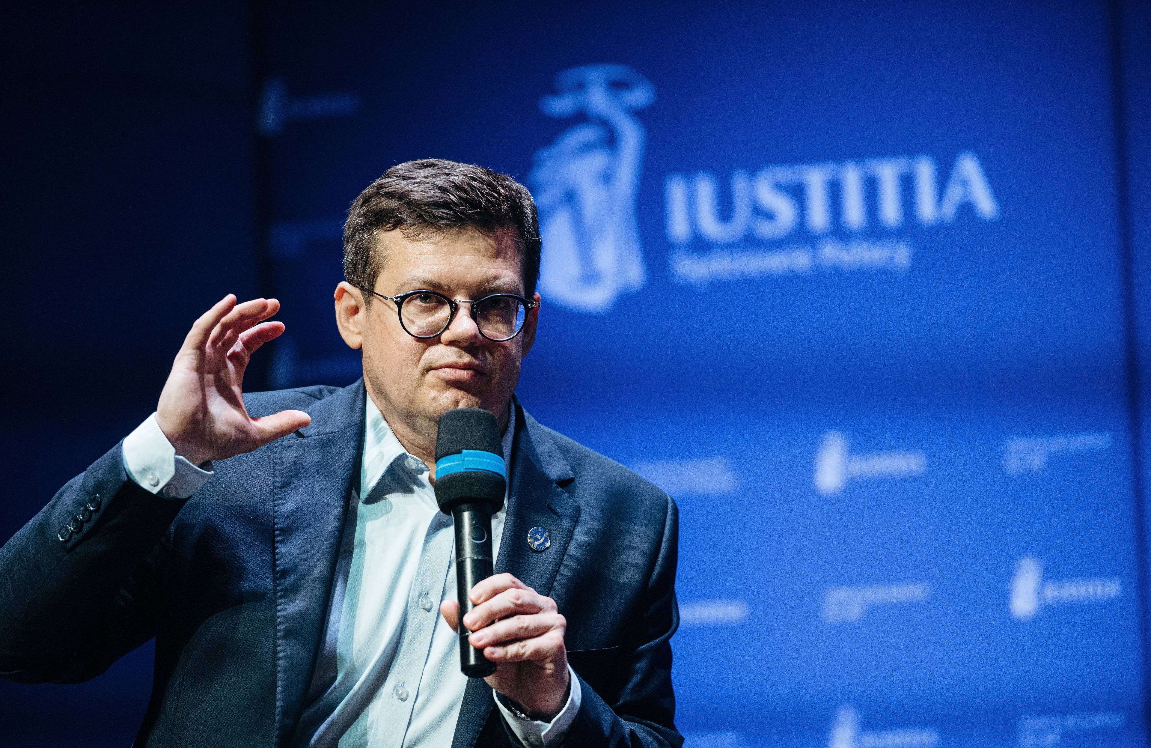 Mężczyzna w okularach przemawia gestykulując na tle napisu Iustitia