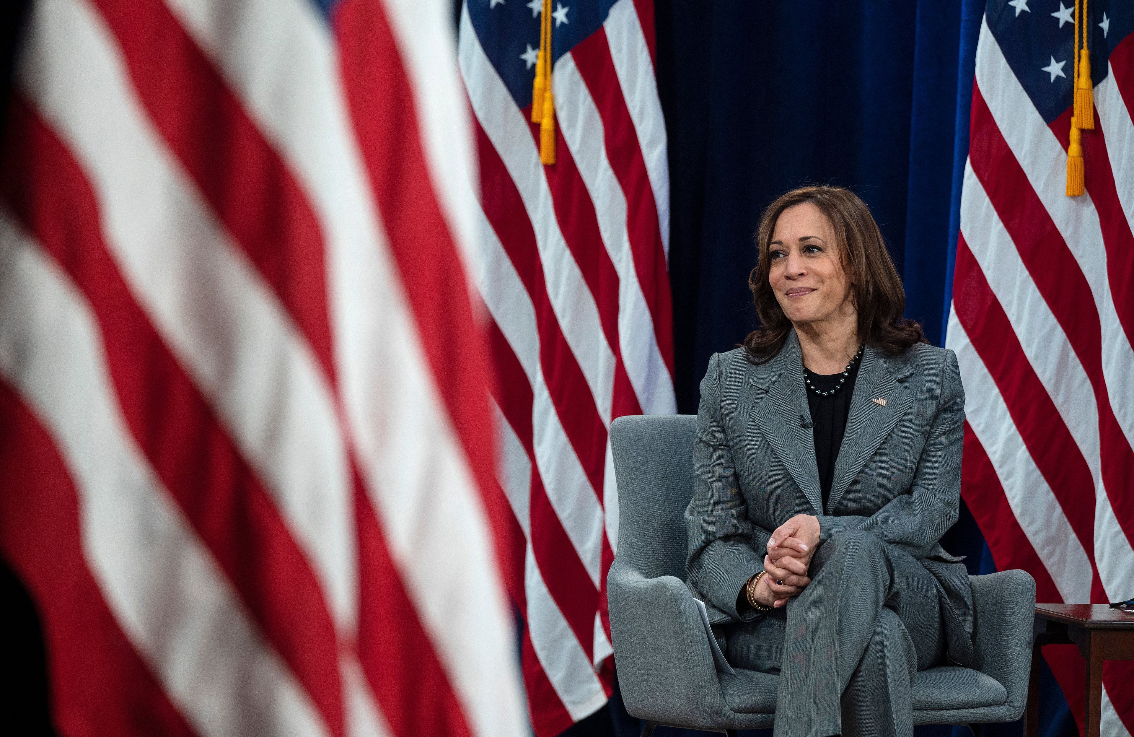Kamala Harris ubrana w szaro-stalowy żakiet siedzi na krześle. Za nią flagi USA