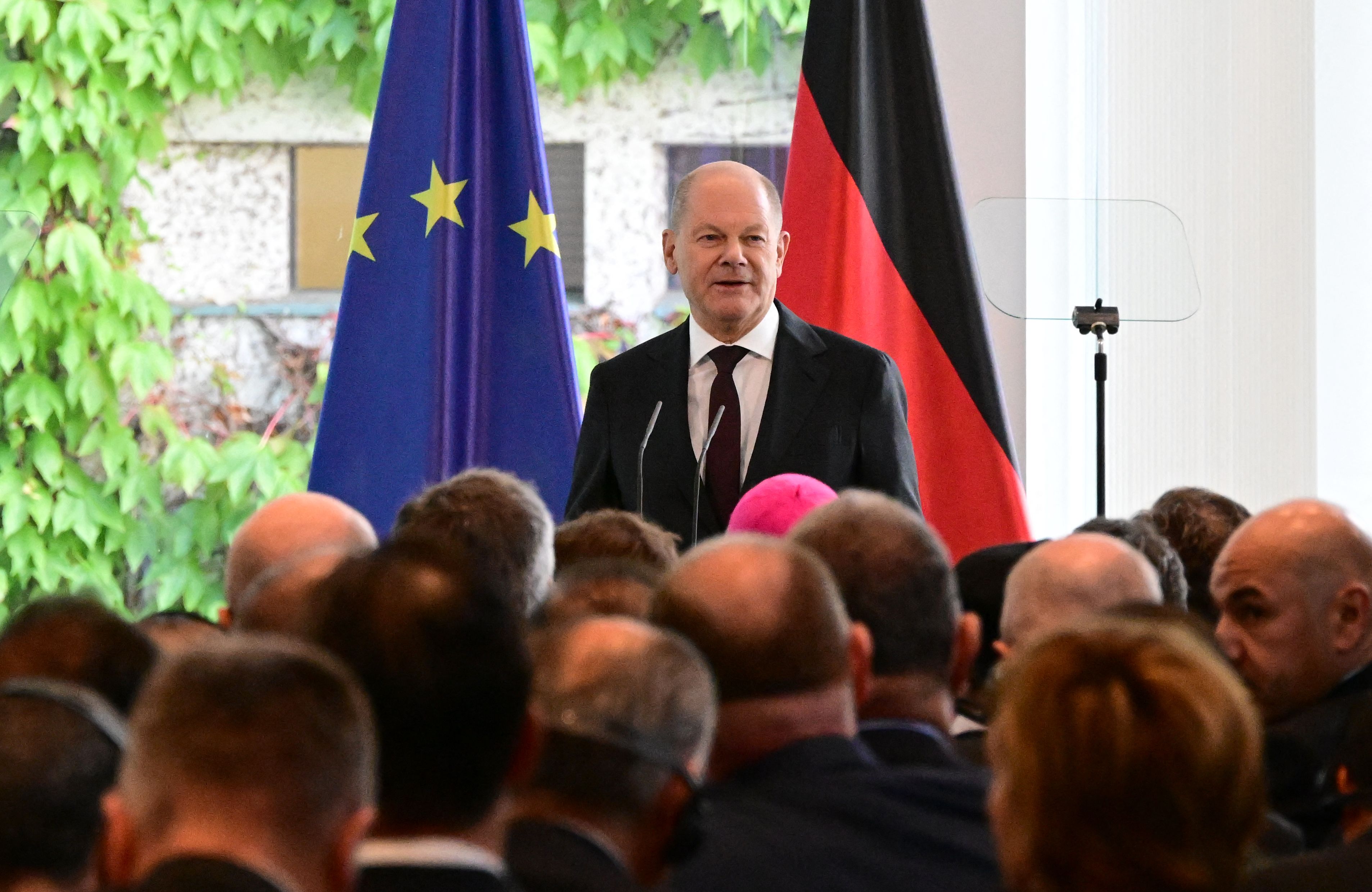 Łysięjący mężczyzna w garniturze stoi na tle flag Niemiec i Unii Europejskiej