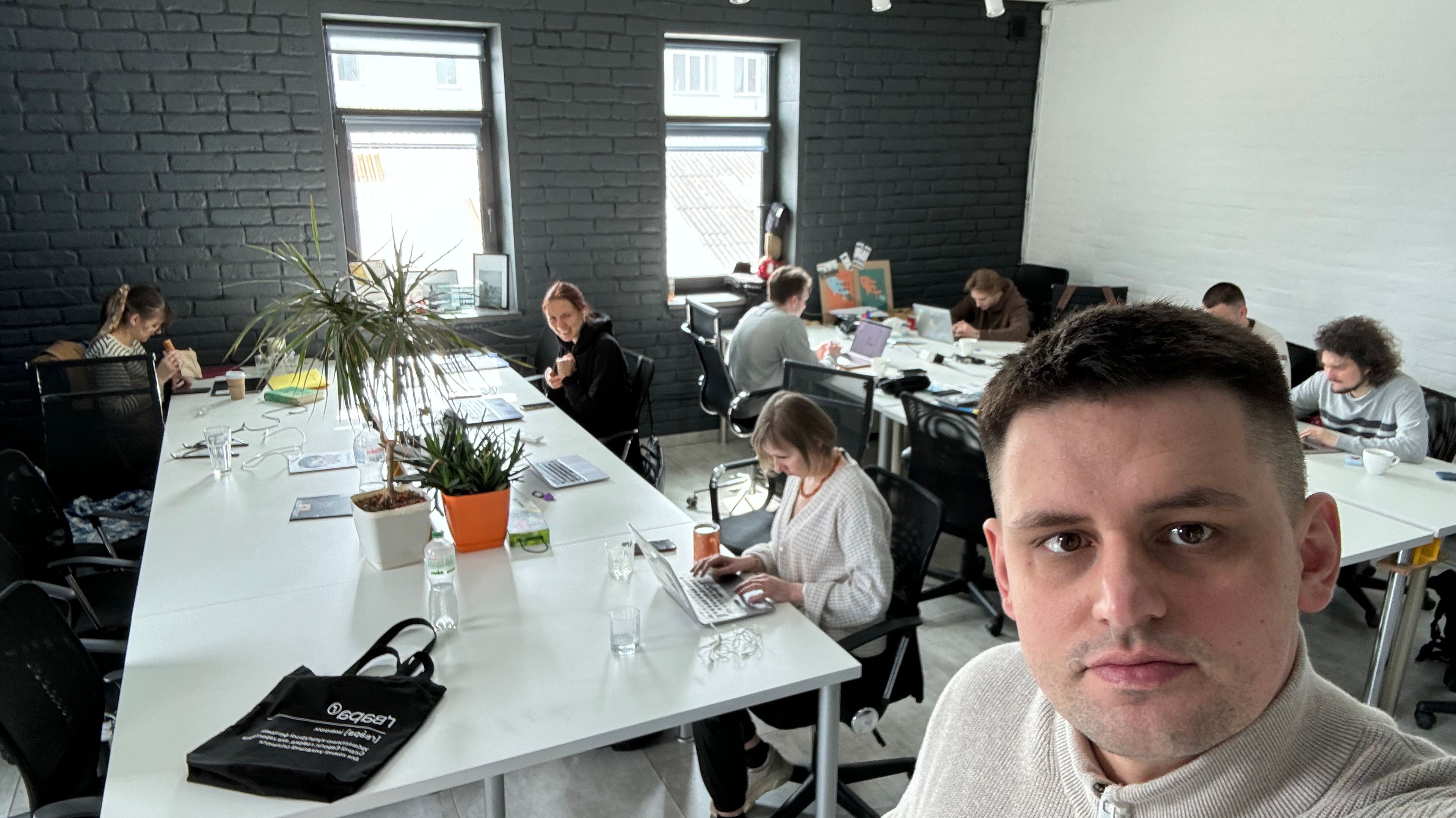 Grupa ludzi, siedzą przy białych biurach, jeden mężczyzna, który robi zdjęcie selfi jest po prawej stronie zdjęcia, jest ubrany w biały sweter