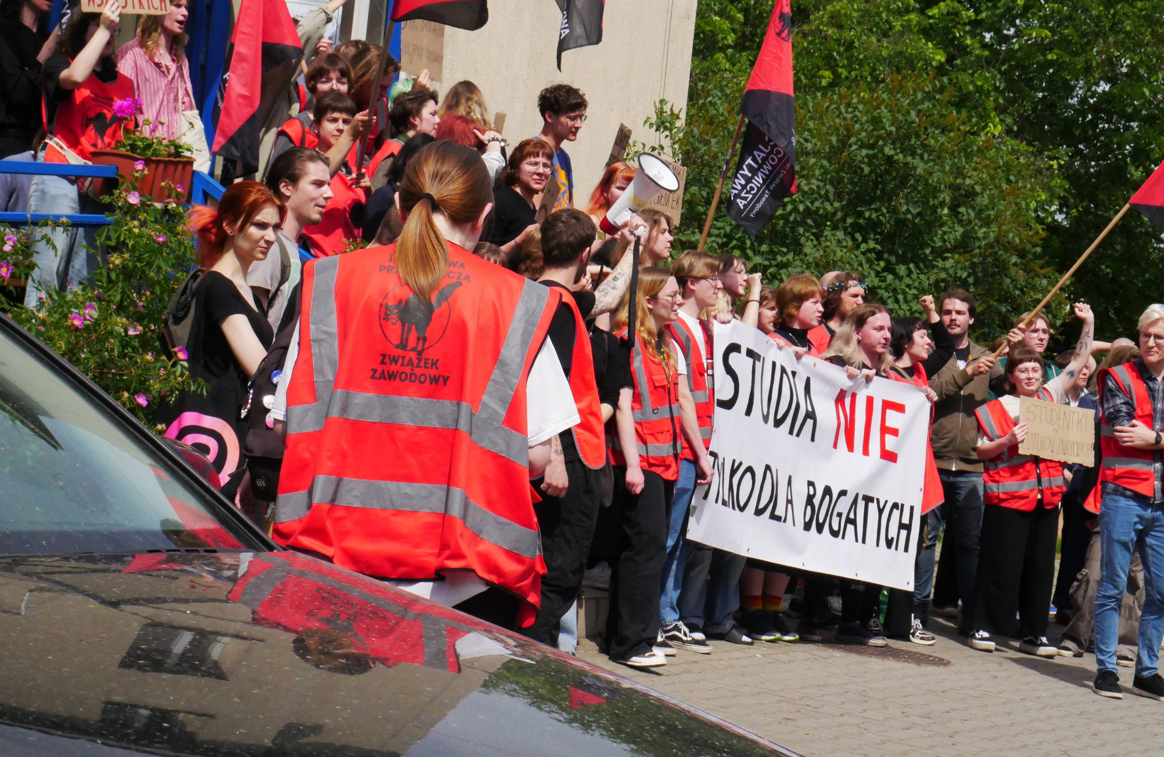 studenci protestujący przed budynkiem akademika w czerwonych kamizelkach. baner z napisem "studia nie tylko dla bogatych"