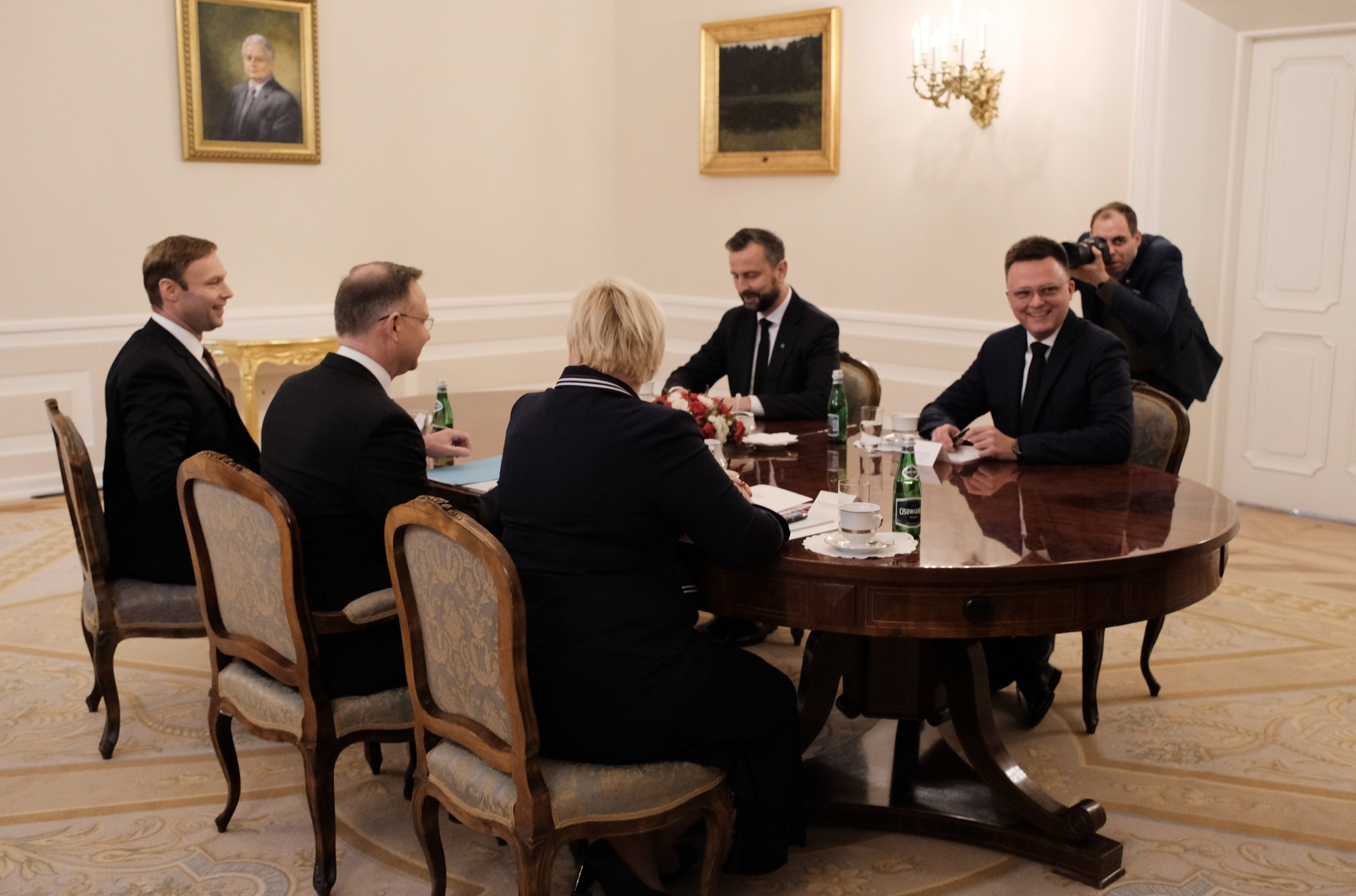 Kilka osób siedzi w ozdobnej sali wokół stołu