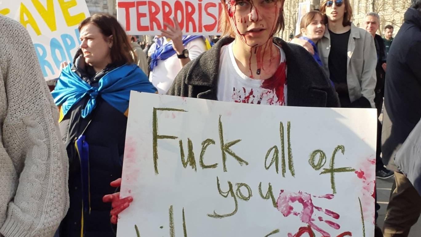 Chłopak z namalowanymi na twarzy śladami krwi trzyma baner z napisem "Fuck all of you with your gas"