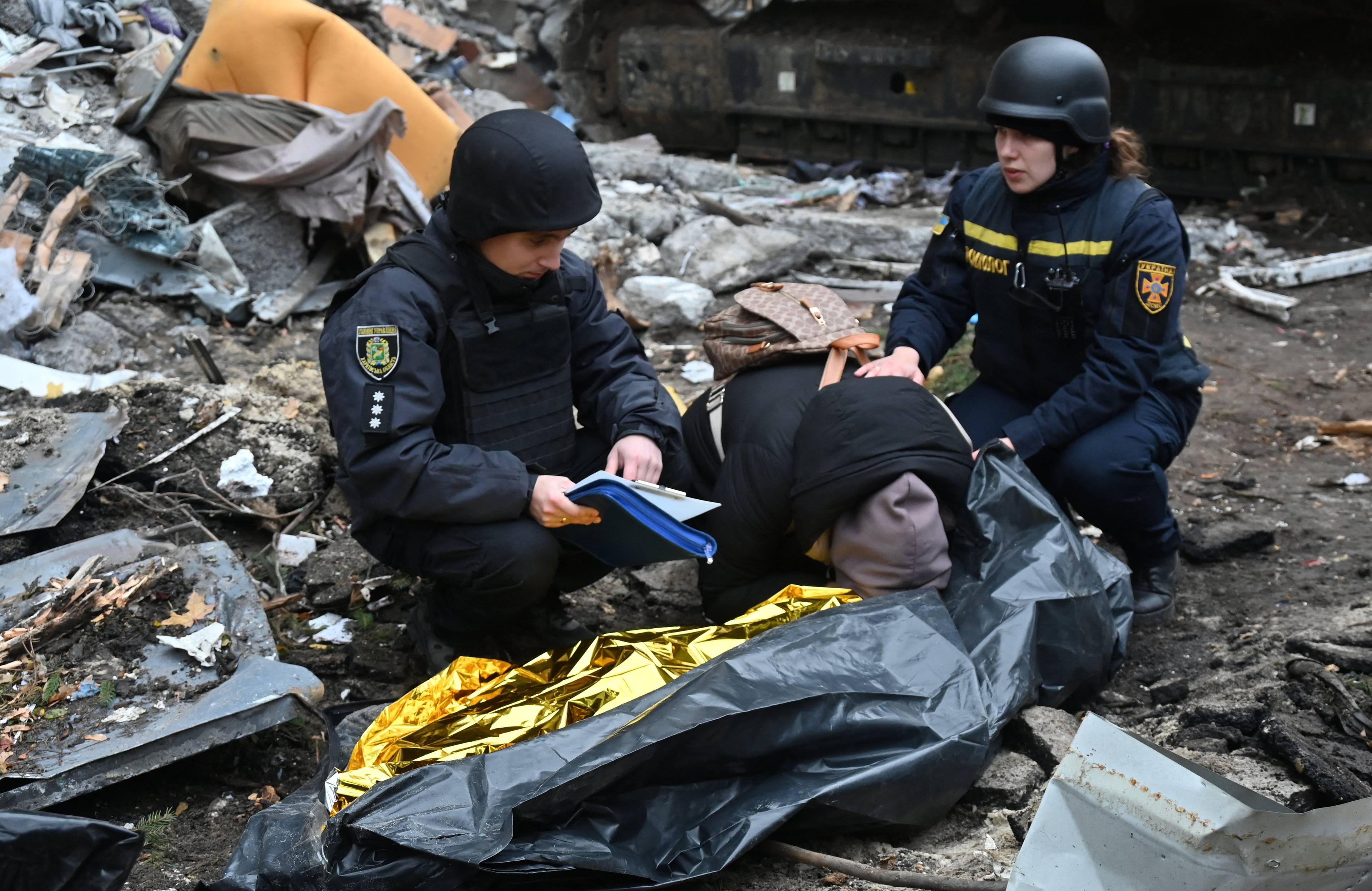 Na zdjęciu są trzy osoby. Przykucnęły wśród gruzów. Kobieta pośrodku, ubrana w czarną kurktkęi ma brązowy kaptur. Pochyliła się nad czarnym workiem z ciałem jej syna.