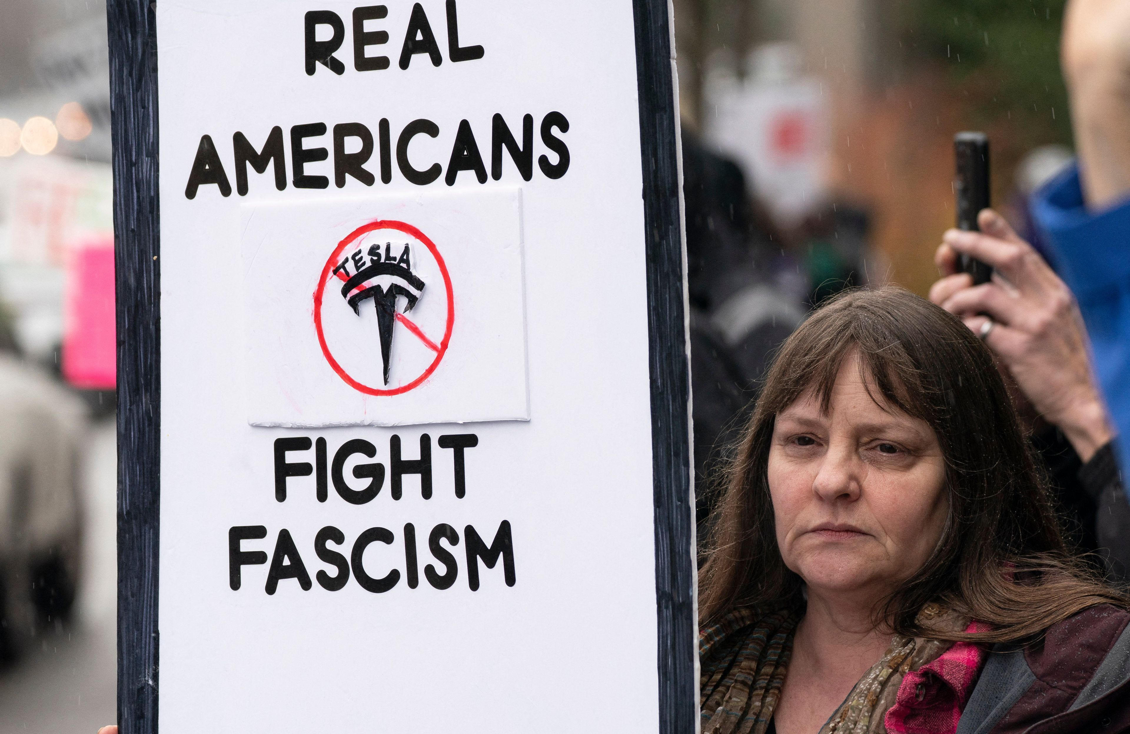 Kobieta trzyma transparent z napisem "Real Americans fight fascism" i przekreślonym logotypem marki Tesla