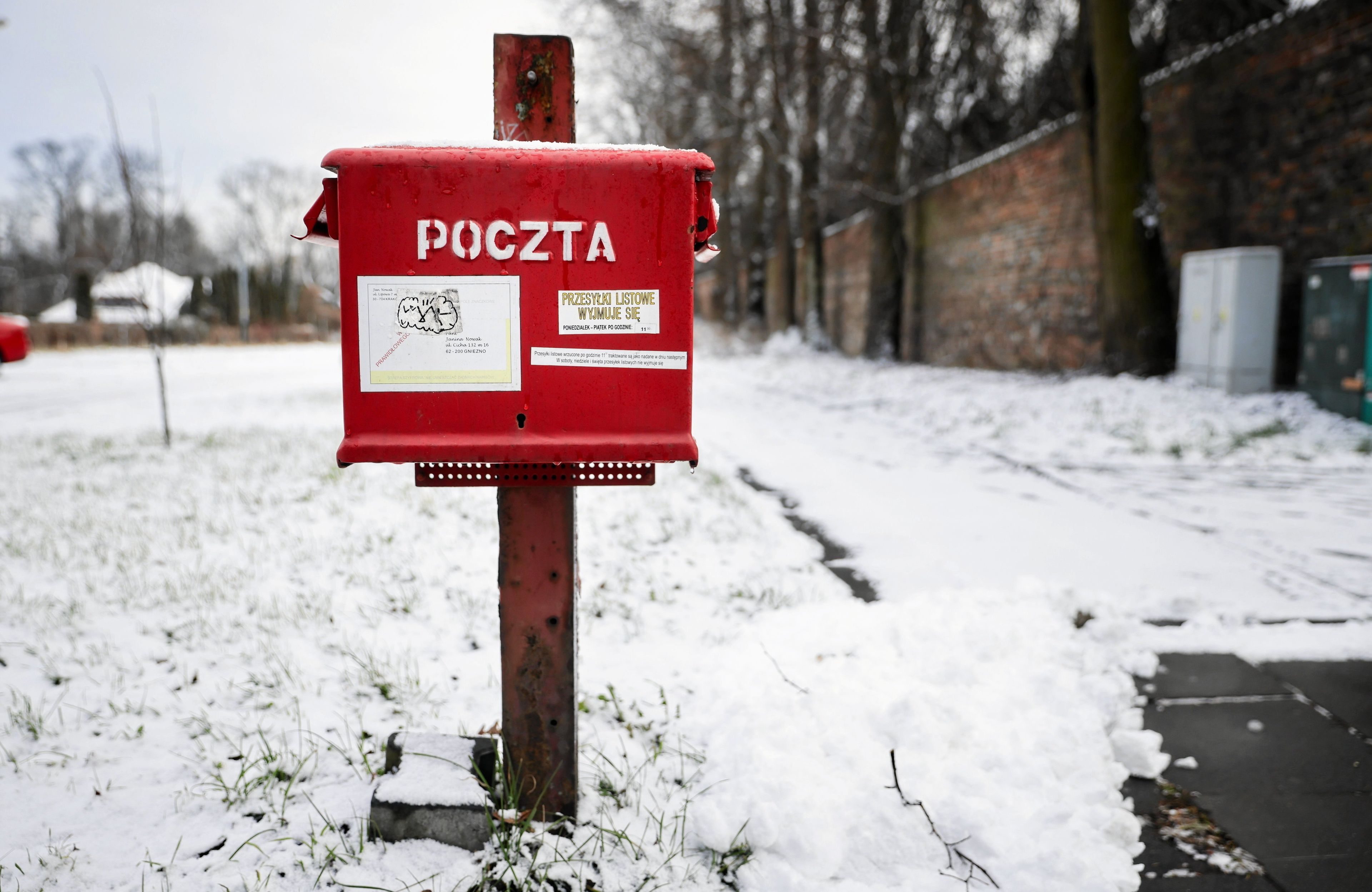 Na zdjęciu widać czerwoną skrzynkę na listy z napisem "POCZTA" stojącą na trawniku pokrytym śniegiem