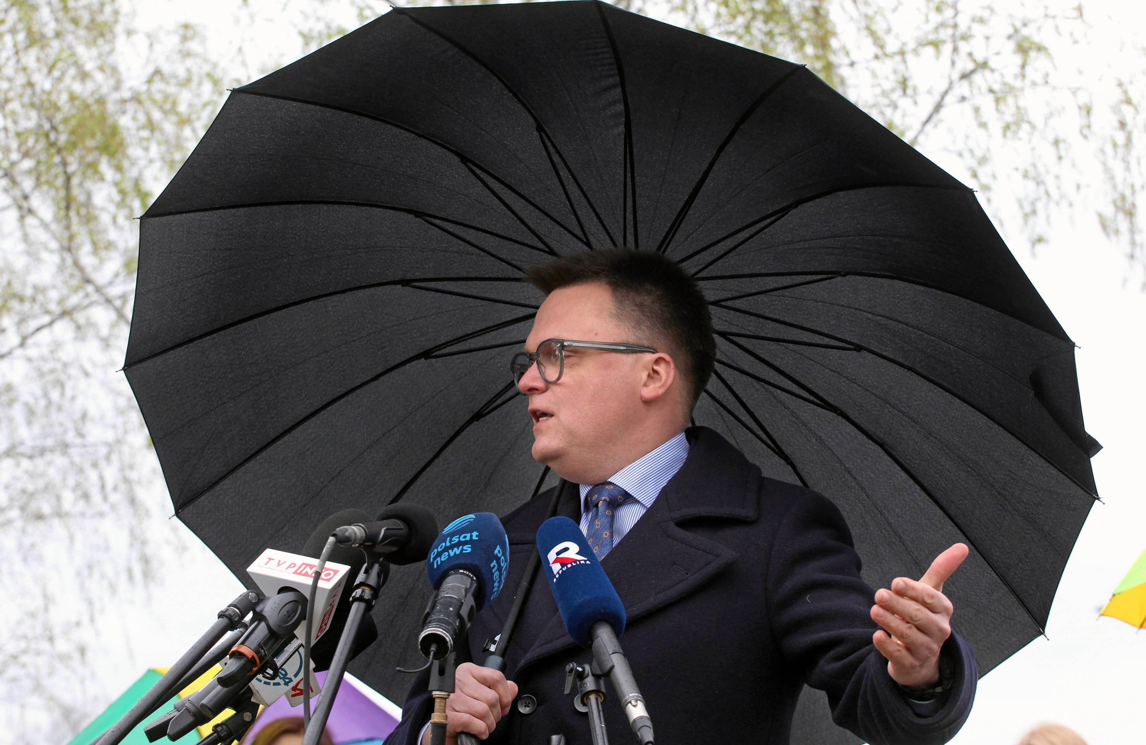 Szymon Hołownia trzyma parasol, przed nim pęk mikrofonów