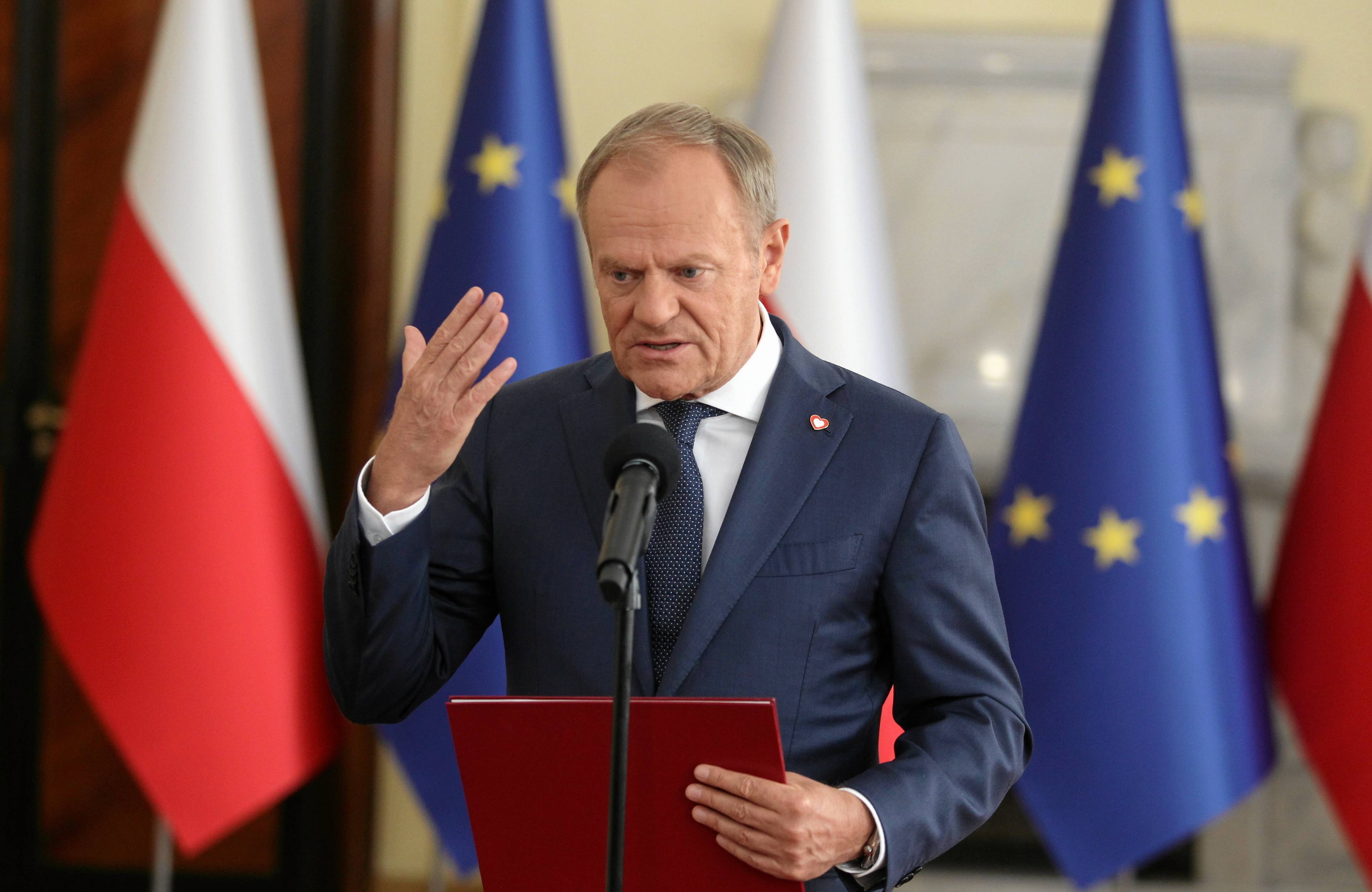 Donald Tusk macha ręką na tle flag Polski i UE stojąc przy mikrofonie