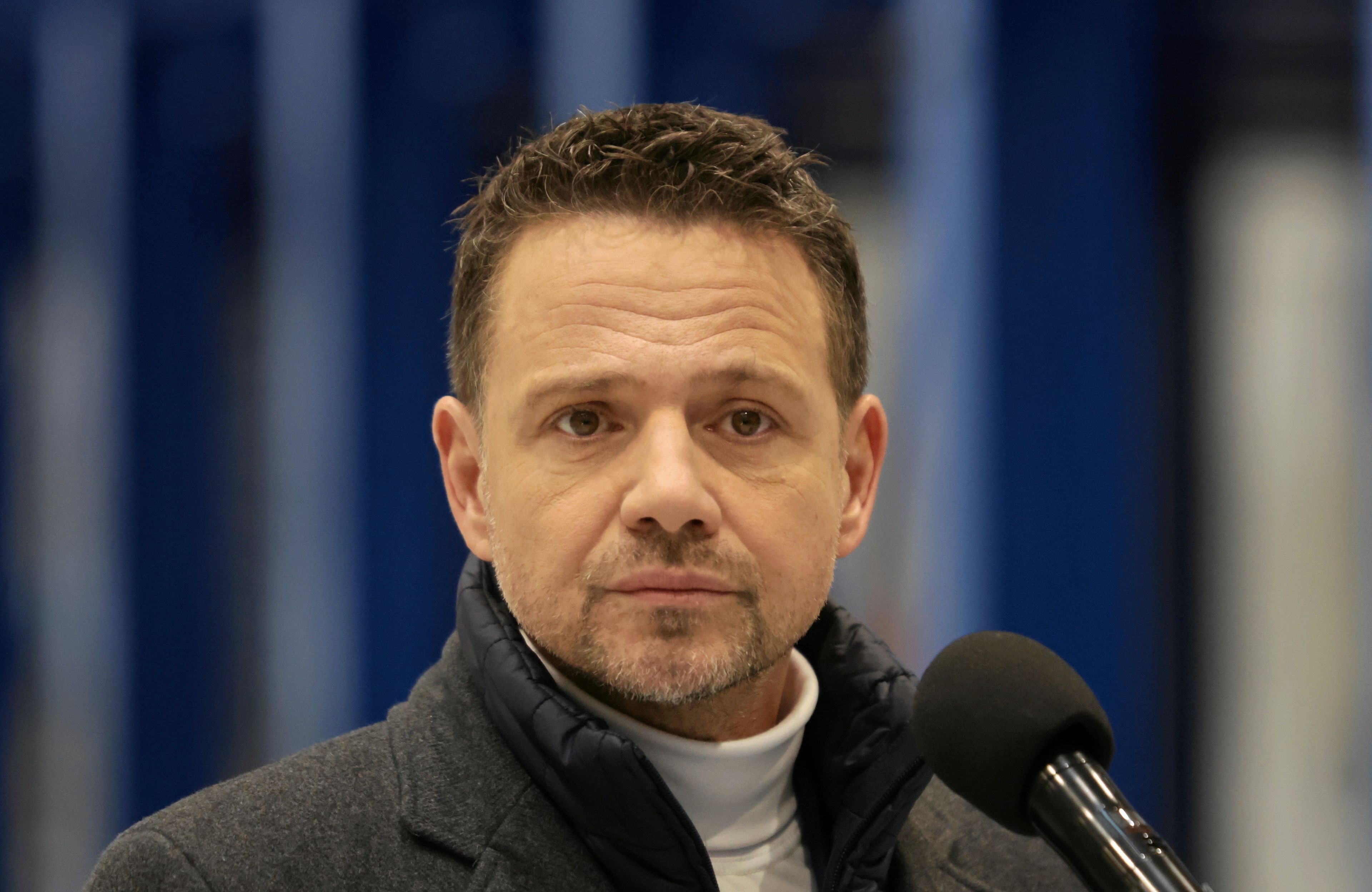 Mężczyzna w białym golfie i ciemnym płaszczu (Rafał Trzaskowski) stoi przed mikrofonem