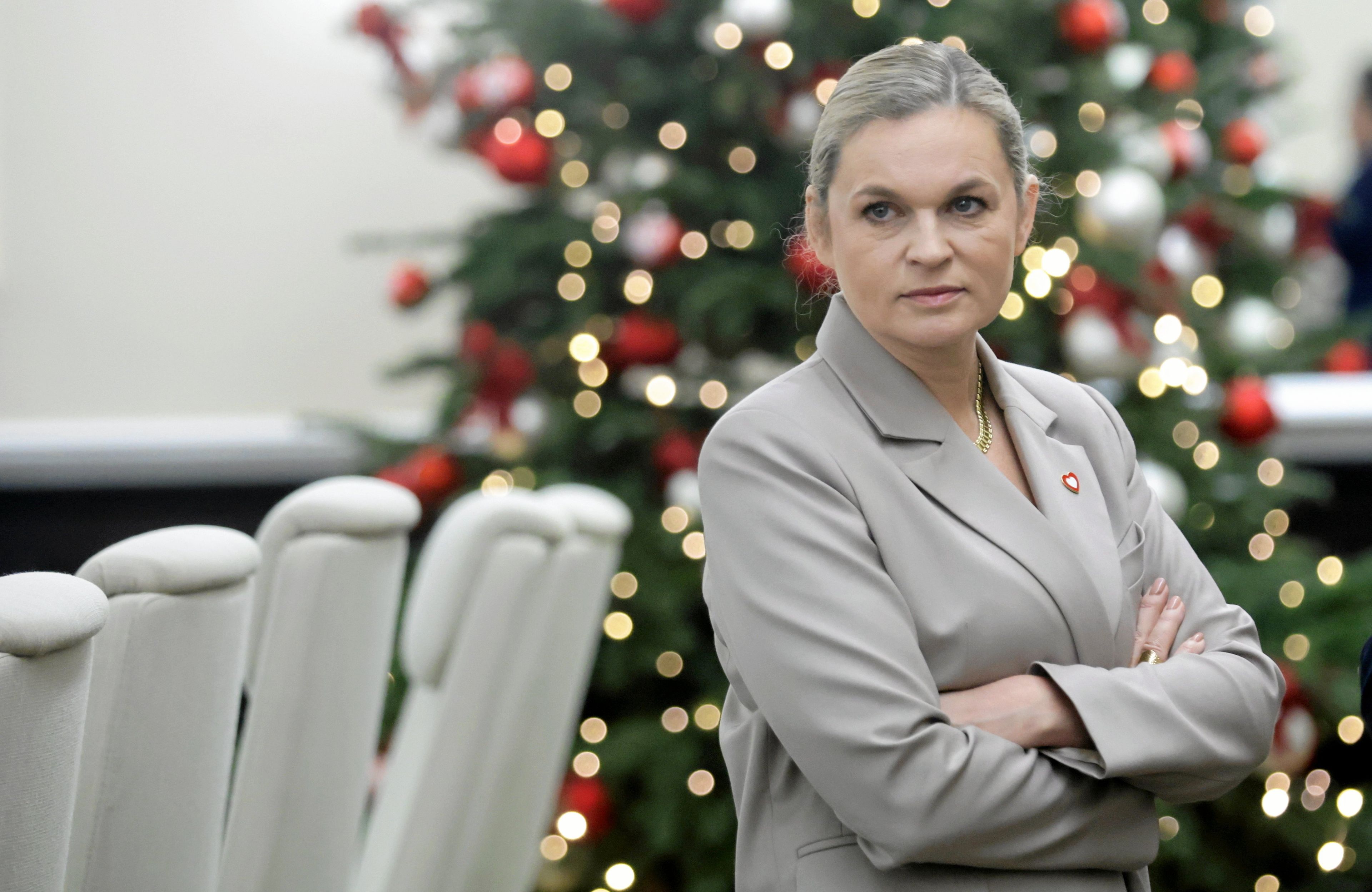 Kobieta w beżowym garniturze (Barbara Nowacka) stoi z założonymi rękami na tle choinki
