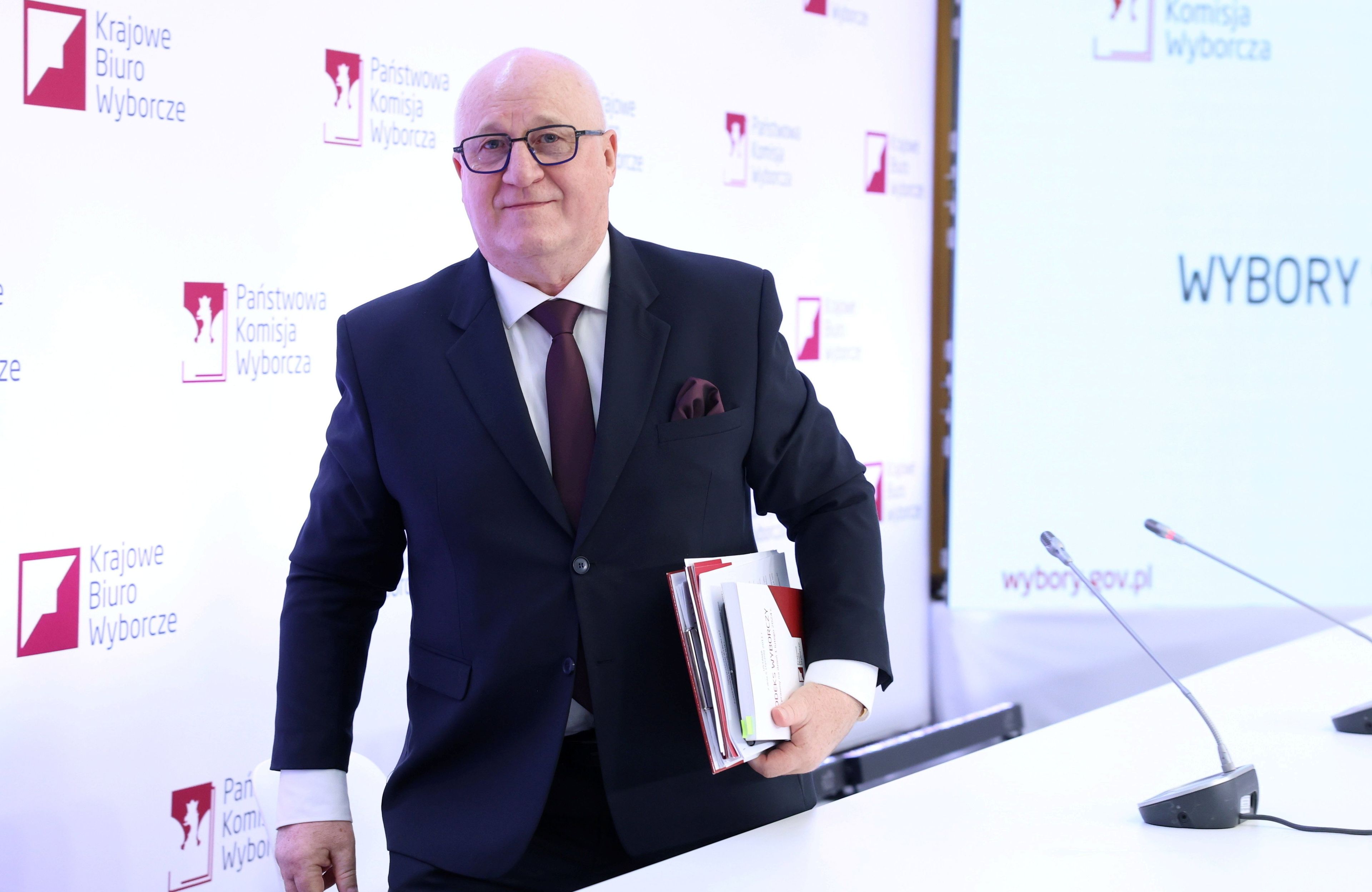 Szef PKW, Sylwester Marcinika, z plikiem papierów pod pachą idzie na tle panelu z logo PKW