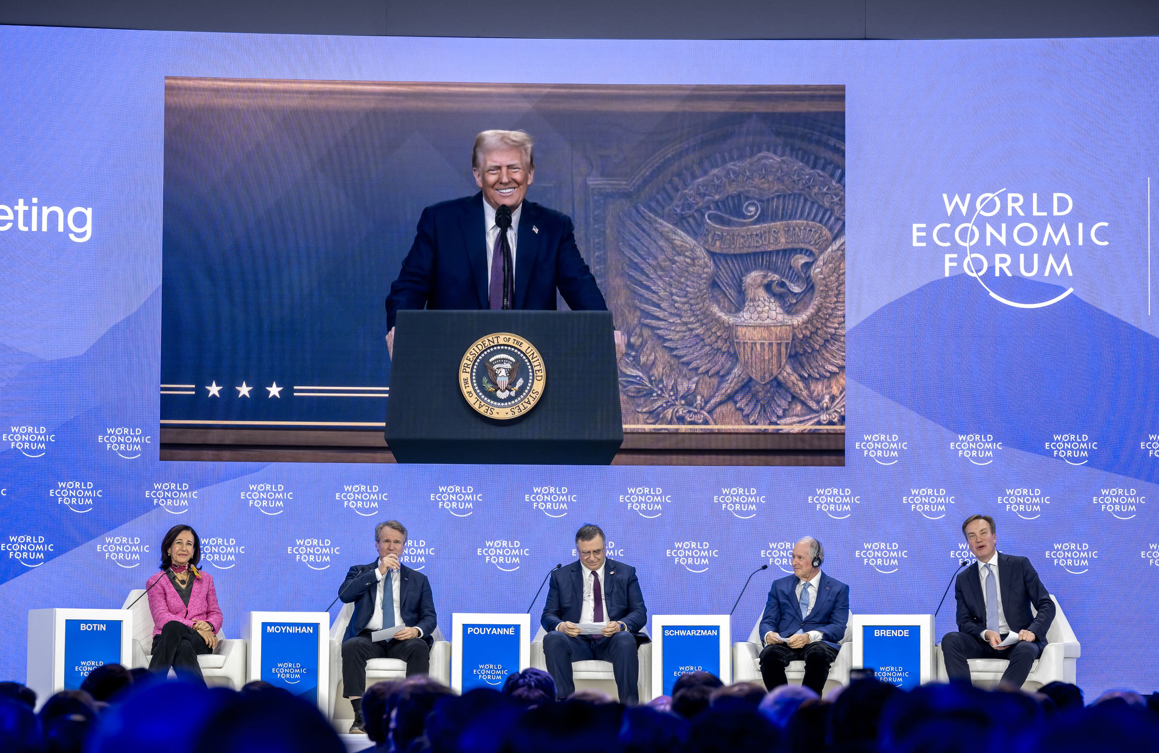Prezydent USA Donald Trump widoczny na wielkim telebimie z szerokim uśmiechem, pod ekranem siedzi kilka osób na fotelach