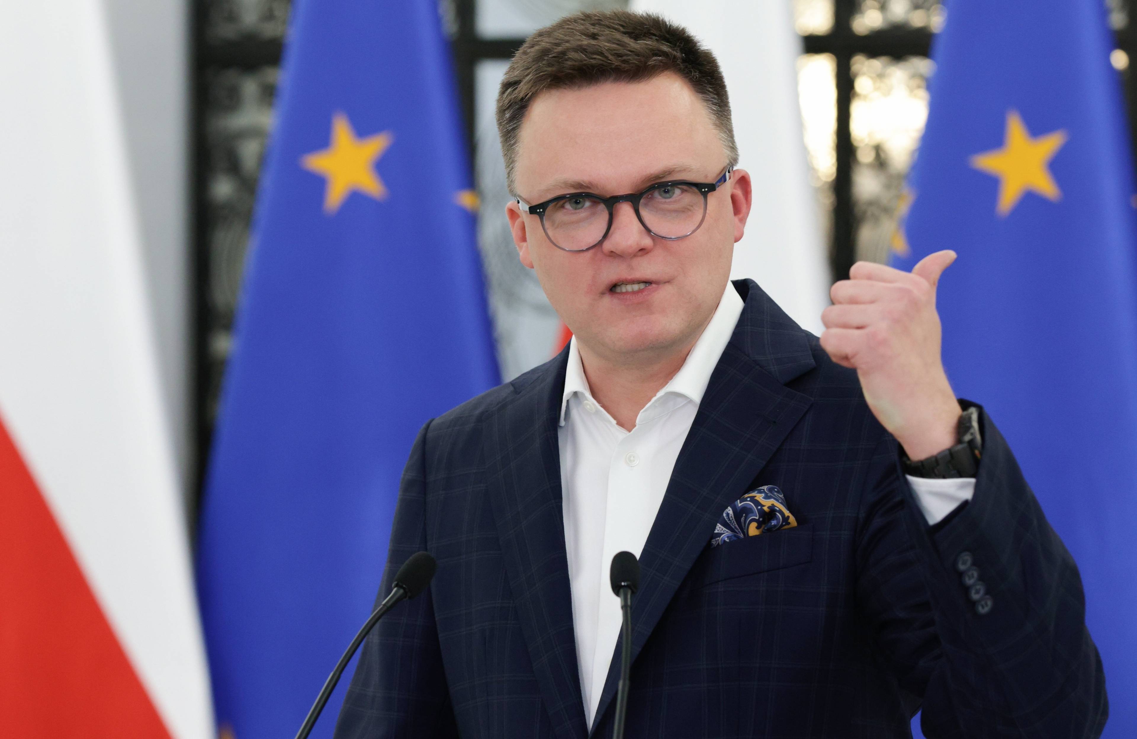 Ostrzyżony na jeża 40-latek w okularach, w garniturze bez krawata, na tle flag Polski i UE