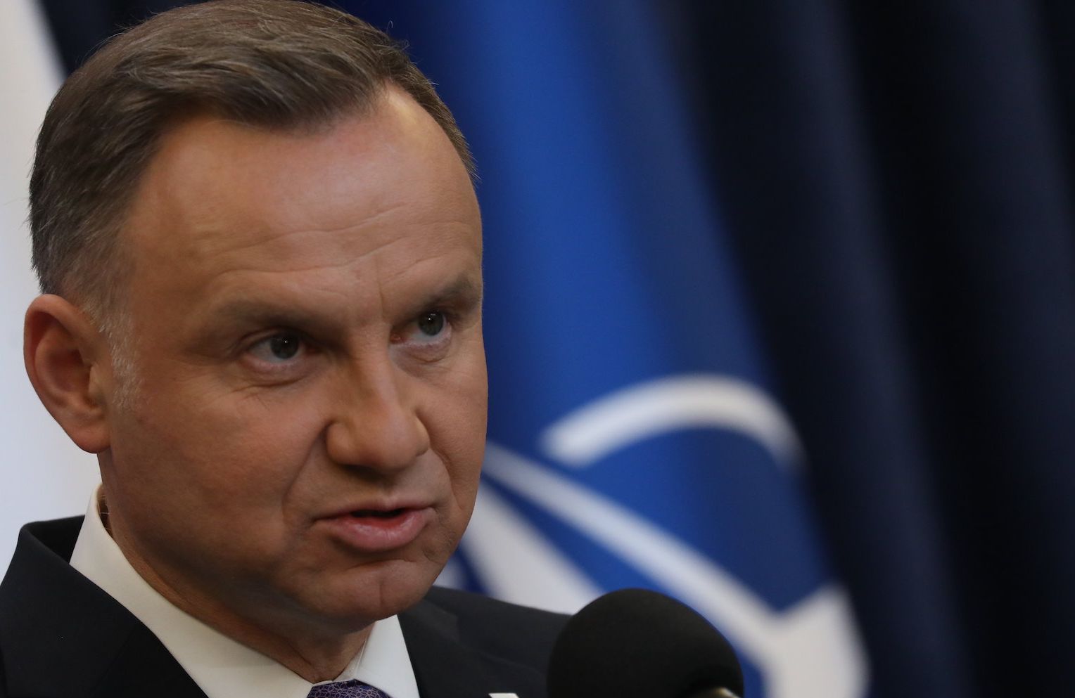 Prezydent Andrzej Duda na tle flag Polski i NATO. Przemawia do mikrofonu z zaciętą miną.