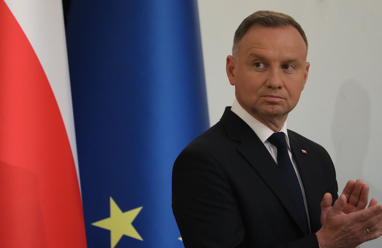 Prezydent Andrzej Duda stoi na tle flagi Polski oraz flagi Unii Europejskiej. Spogląda w prawo z poważną miną i bije brawo.