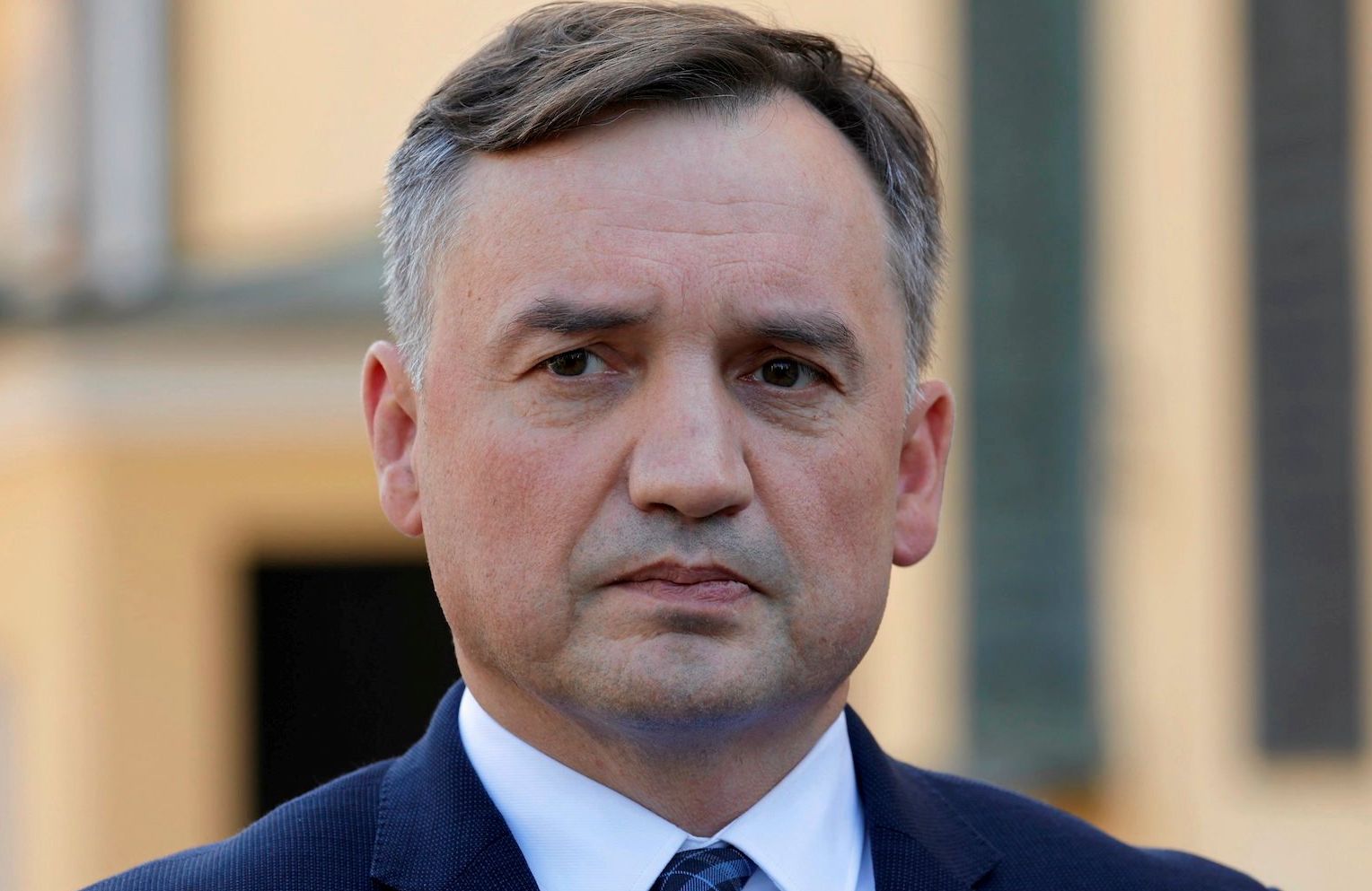 Zbigniew Ziobro w granatowym garniturze, białej koszuli lekko przekrzywionym krawacie w ganatowo-niebieską kratkę. Patrzy w bok z zafrasowaną miną