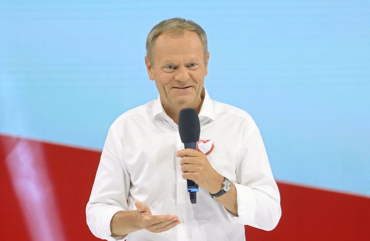 Donald Tusk na konwencji PO w Tarnowie. Przemawia na tle biało czerwonej flagi ubrany w białą koszulę i granatowe spodnie.