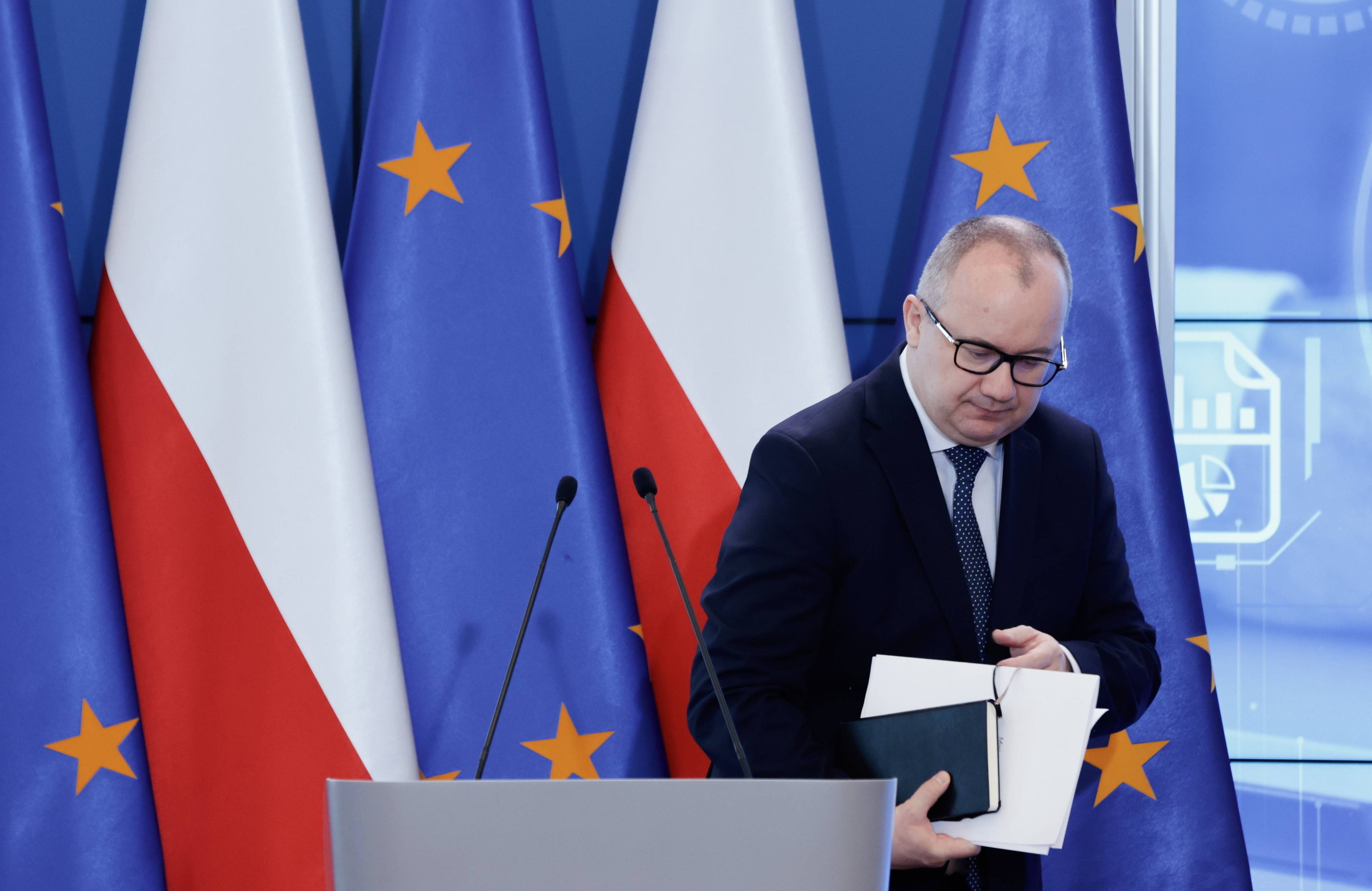 Adam Bodnar schodzi z mównicy, w ręce trzyma notes i papiery, za nim flagi Polski i UE