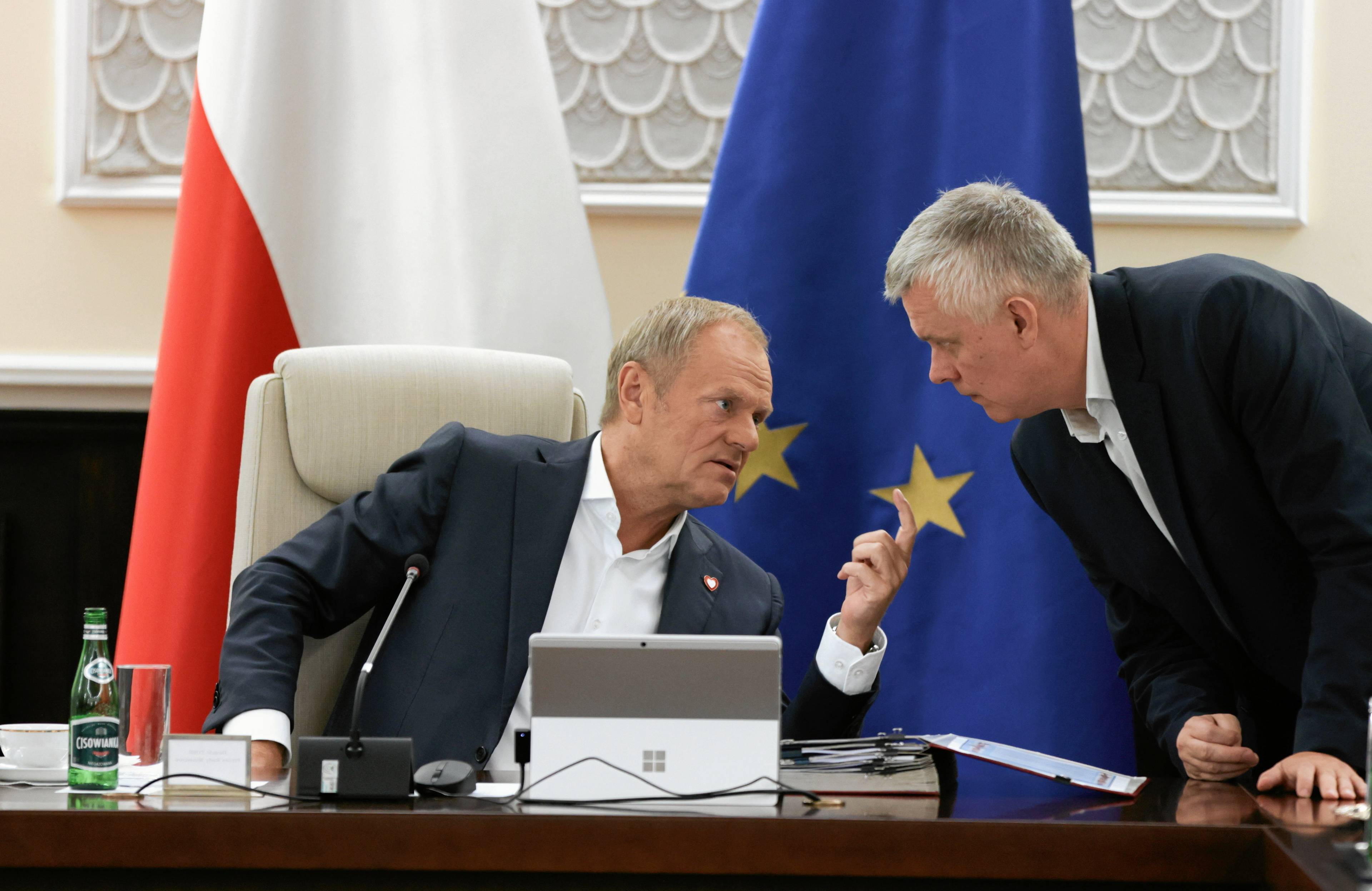 Premier Donald Tusk (z lewej) na fotelu, za nim flaga Polski i Unii Europejskiej. Obok niego, z prawej, minister Tomasz Siemoniak. Nachyla się do premiera.