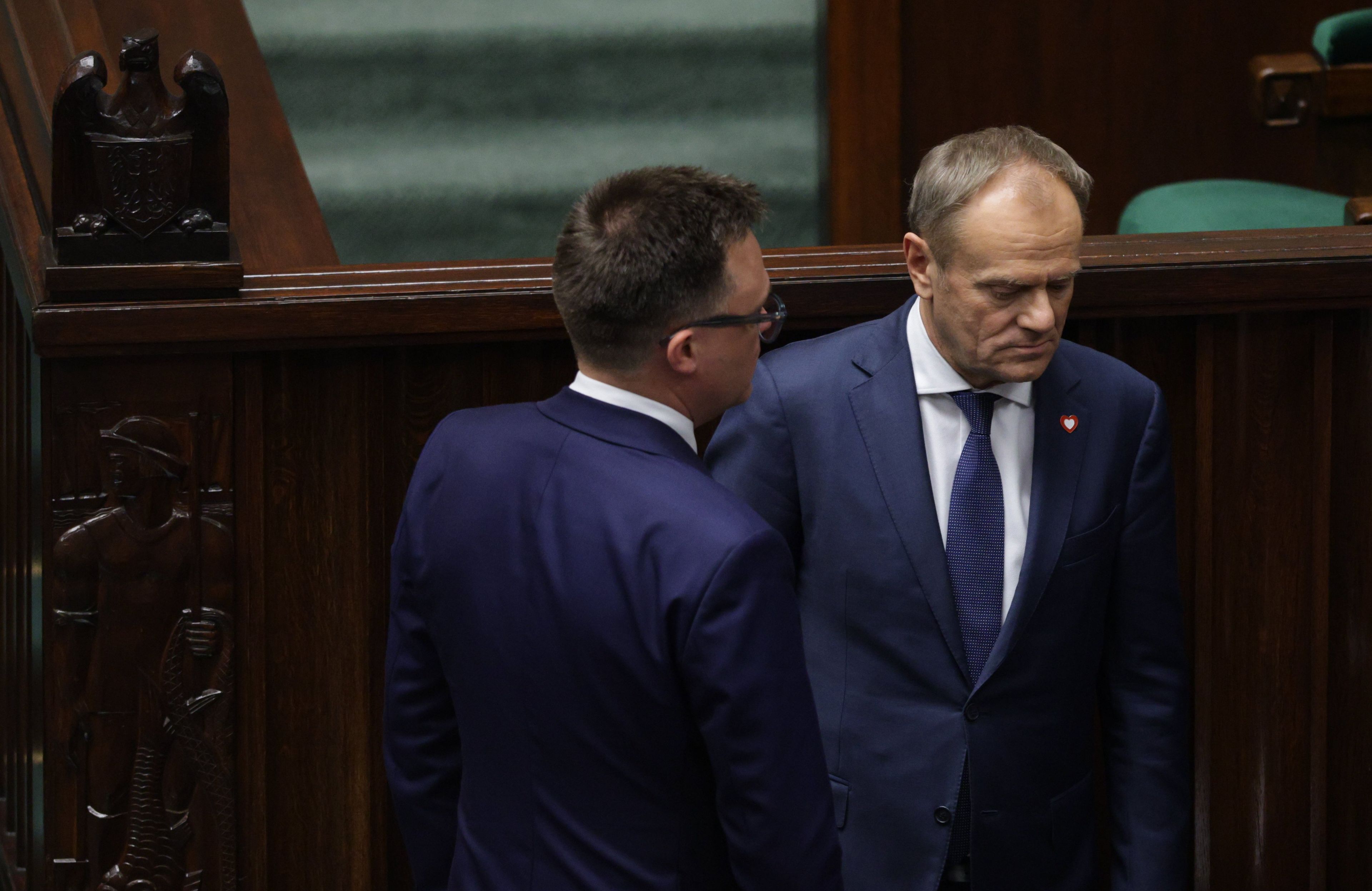 Dwóch mężczyzn, jeden odwrócony tyłem, drugi poważny i smutny, wokół drewniane meble
