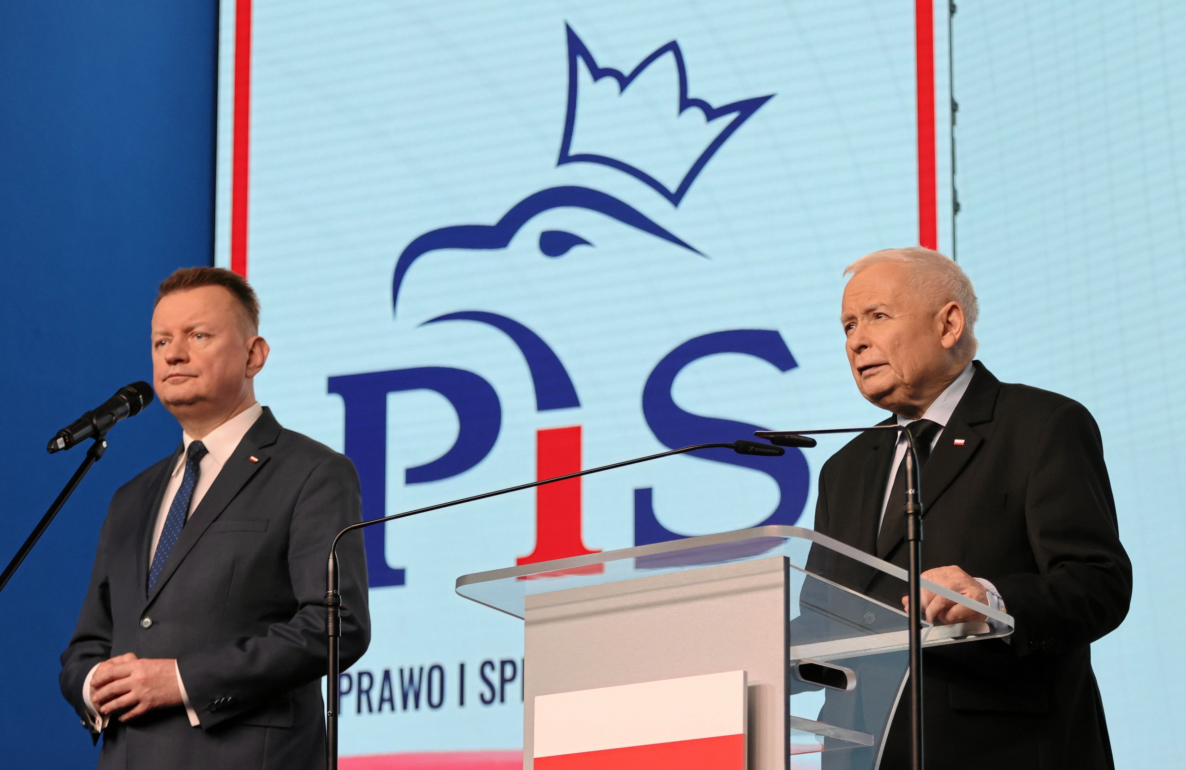 Dwóch mężczyzn, jeden za szklanym pulpitem z biało-czerwoną flagą. Za nimi logo PiS z orłem w koronie. Mariusz Błaszczak i Jarosław Kaczyński